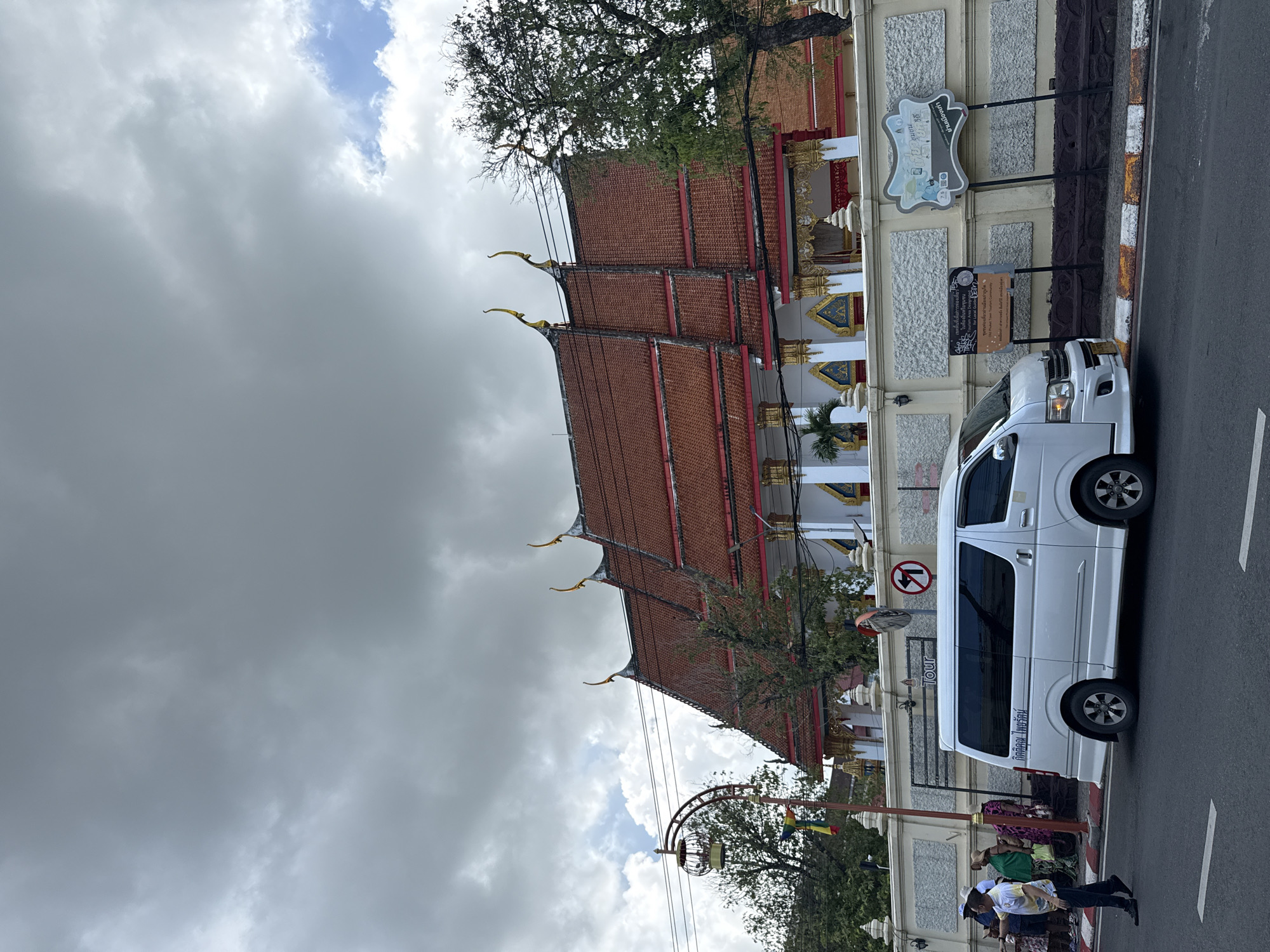 The image depicts a street scene in Phuket City Municipality, Thailand. In the foreground, a white van is parked on the side of the road, with the word "Tour" visible on its side. Several people are standing near the van. Behind the van, a traditional Thai temple with a red tiled roof and ornate golden embellishments is visible. The temple building features a long facade with decorative columns and windows.
The sky is heavily overcast, suggesting a cloudy or potentially rainy day. A traffic sign with a red circle and a diagonal line through it, indicating "no entry" or a similar restriction, is mounted on a pole in front of the temple. Another sign with Thai script and some graphic elements is also present. Trees with lush green foliage are interspersed between the temple and the road. The overall atmosphere appears to be a typical day in a bustling urban environment with cultural landmarks. The time of day is not explicitly indicated but the lighting suggests it is daytime.