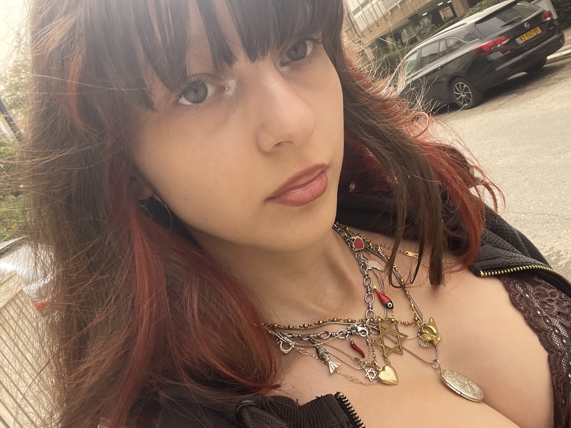 A woman with dark brown hair with red highlights and bangs is pictured in a close-up selfie. She has light eyes, full lips with gloss, and is wearing multiple layered necklaces with various pendants including a Star of David and a heart. She is wearing a dark, possibly brown, jacket with a visible zipper and a lace-trimmed top underneath. In the background, out of focus, is a dark-colored station wagon with a license plate that appears to read "83-640-55". Buildings are also visible in the distance. The location is identifiable as Petah Tikva, Israel, based on the license plate format.