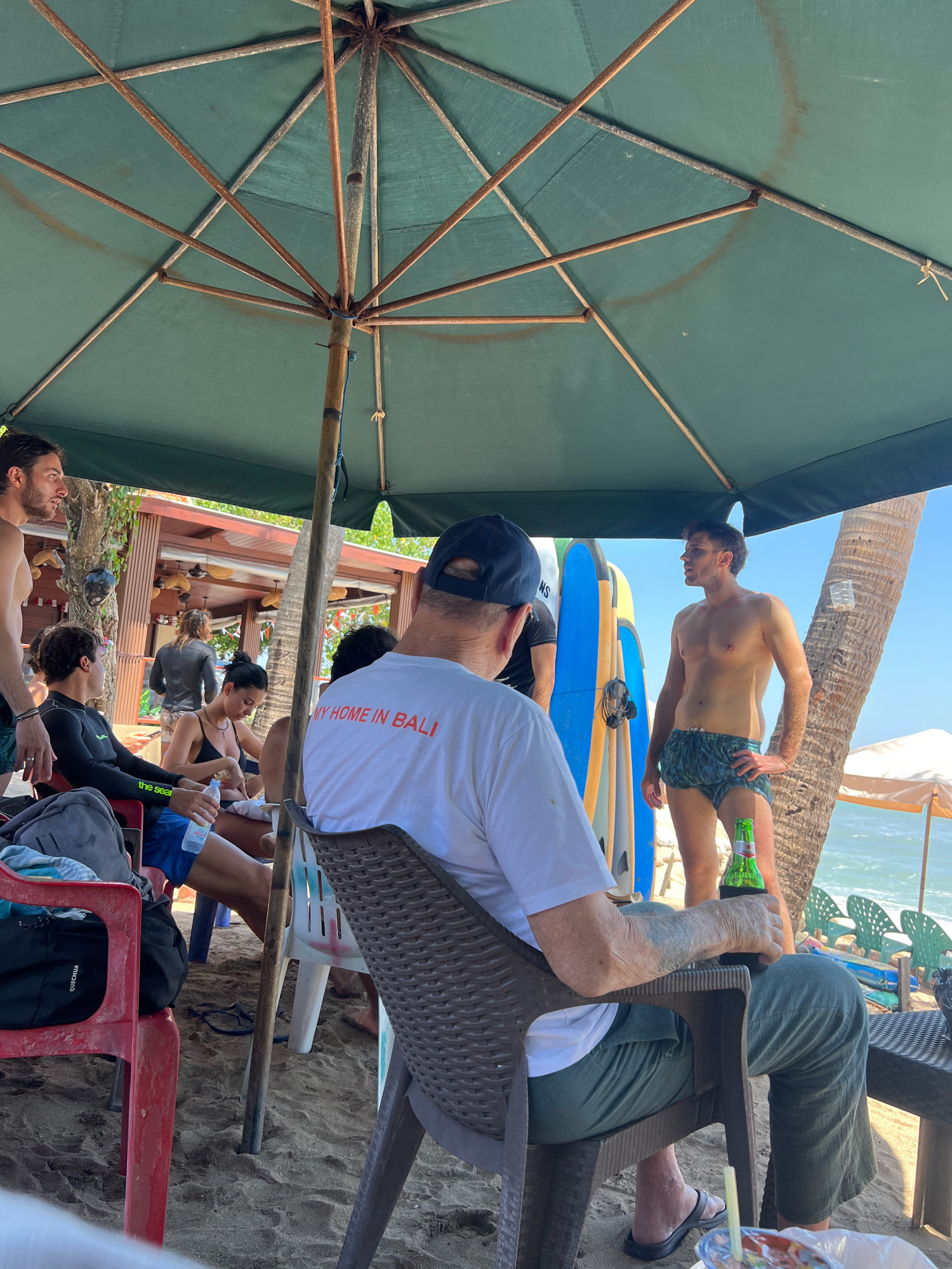 The image depicts a group of people at a beachside establishment in Kuta, Indonesia, likely a bar or restaurant. A large green umbrella provides shade for a group of individuals seated on the sand. In the foreground, a man wearing a white t-shirt with the text "MY HOME IN BALI" printed in red, sits in a brown plastic chair, holding a beverage in a koozie. To his left, another man in blue shorts and a black wetsuit sits with a water bottle. A woman in a black bikini is seated beside him. In the background, a shirtless man in patterned shorts stands, holding a green beer bottle. Behind him, surfboards are propped up against a structure. The environment is sandy, with palm trees and the ocean visible in the distance. The overall atmosphere suggests a relaxed beach scene.