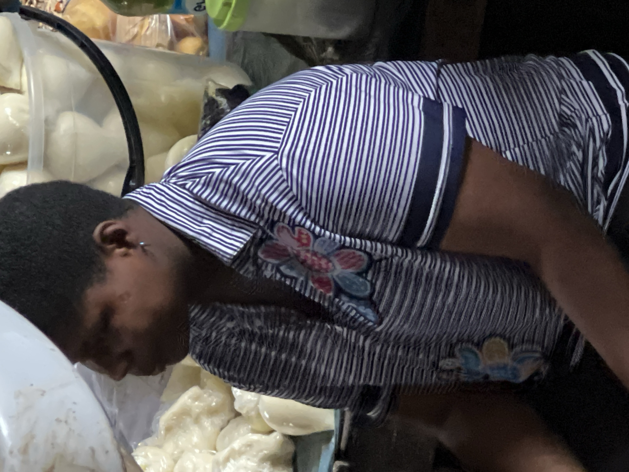 The image captures a close-up view of a person wearing a blue and white striped shirt with a floral applique on the left side of the chest. The person has dark skin and short, dark hair, and is wearing a small silver earring. In the background, a clear plastic bucket is filled with white, rounded objects, possibly food items like dumplings or buns. The setting appears to be a market or a food stall, with other indistinct objects and containers visible in the periphery. The lighting suggests an indoor environment, possibly with overhead artificial light.