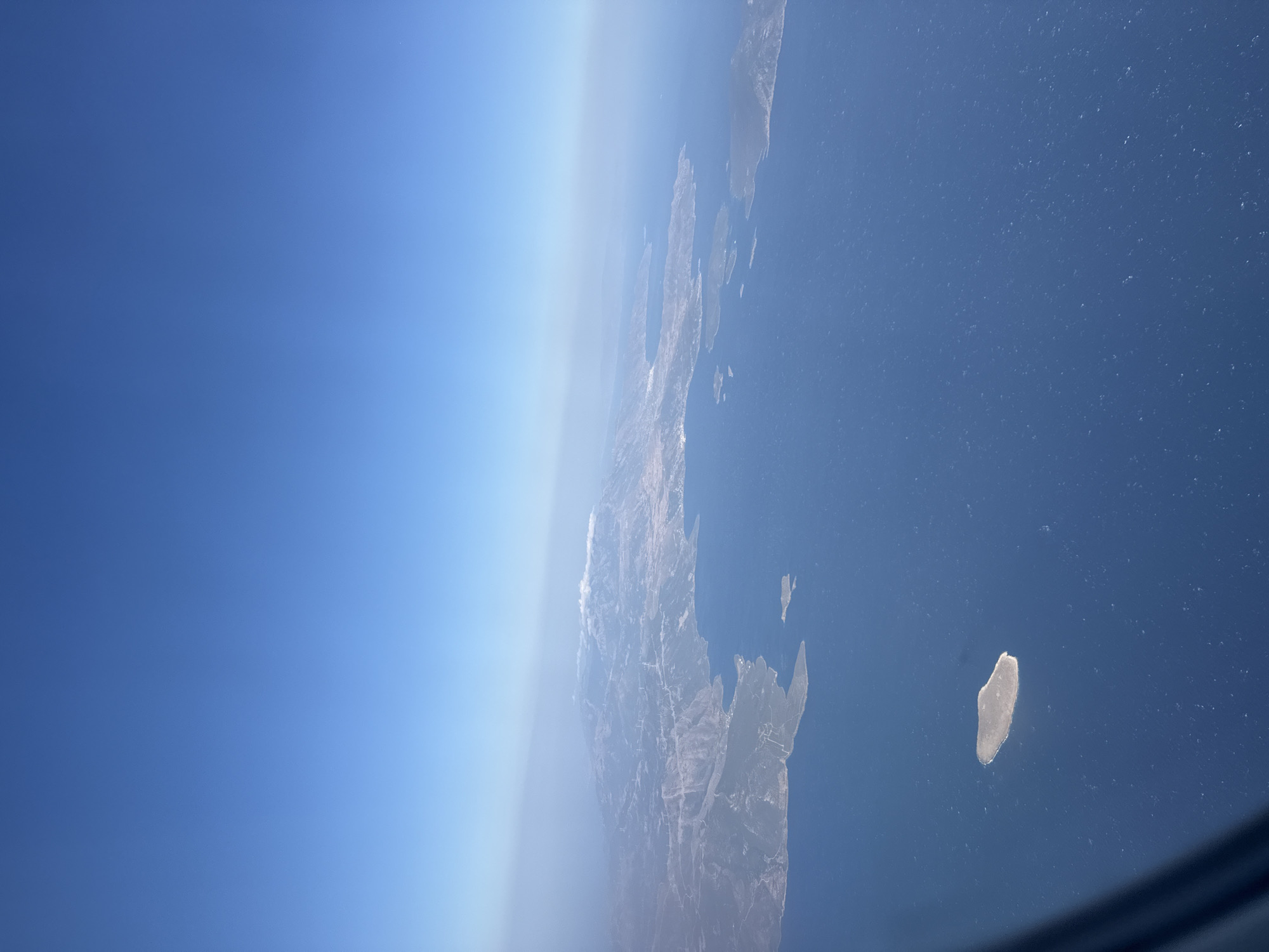 An aerial view captures a vast expanse of deep blue ocean dotted with whitecaps, meeting a rugged coastline with sparse vegetation. Several islands of varying sizes are scattered across the water, some appearing rocky and barren while others show signs of green. The mainland features a mountainous terrain, with a hazy layer of atmosphere obscuring the distant peaks. Wind turbines are visible on some of the hillsides, suggesting human presence and development. The sky is a clear, pale blue, transitioning to a lighter hue near the horizon. The perspective suggests the image was taken from an airplane window, with a dark shadow or portion of the aircraft visible in the lower left corner. The scene appears to be a coastal region in Greece, characterized by its islands and mountainous topography.