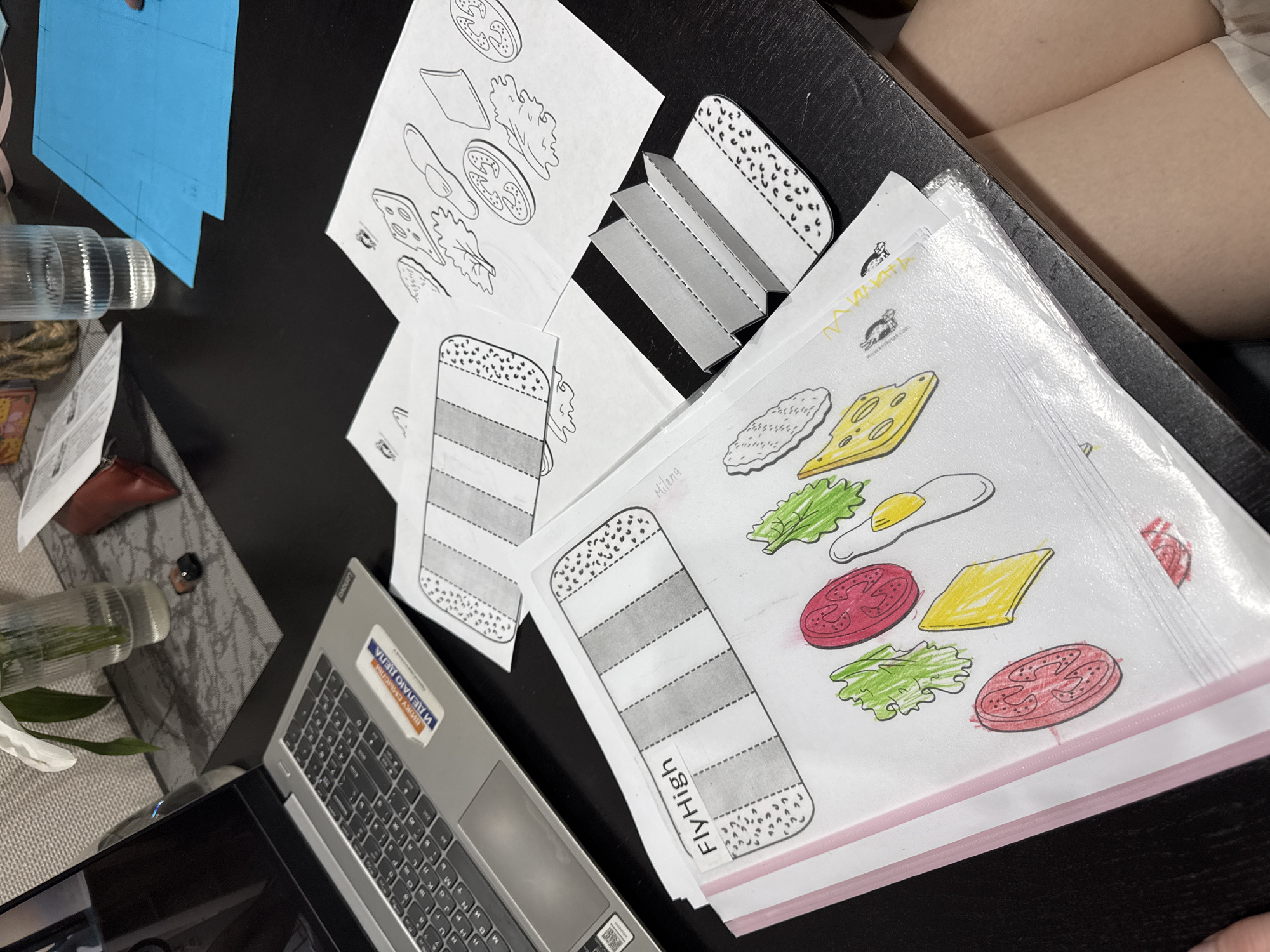 The scene depicts a dark-colored desk surface cluttered with various items. Predominantly, there are printed papers featuring illustrations of sandwich components like bread slices, lettuce, cheese, tomatoes, and eggs. Some of the papers are colored, while others are in black and white. There's a partially visible laptop to the lower left of the frame. To the left of the laptop, a glass container and other small items are present. A blue, folded paper is visible in the top left corner. A fair-skinned person's leg is seen in the upper right corner. The sticker on the laptop keyboard indicates the Cyrillic alphabet. The name "Milena" is visible on one of the papers. The text "FIVHIGh" is printed on the bottom of one of the bread slice illustrations. While no definitive visual cues confirm the city of Perm, Russia, the presence of the Cyrillic alphabet suggests a location within a region where Russian is used.