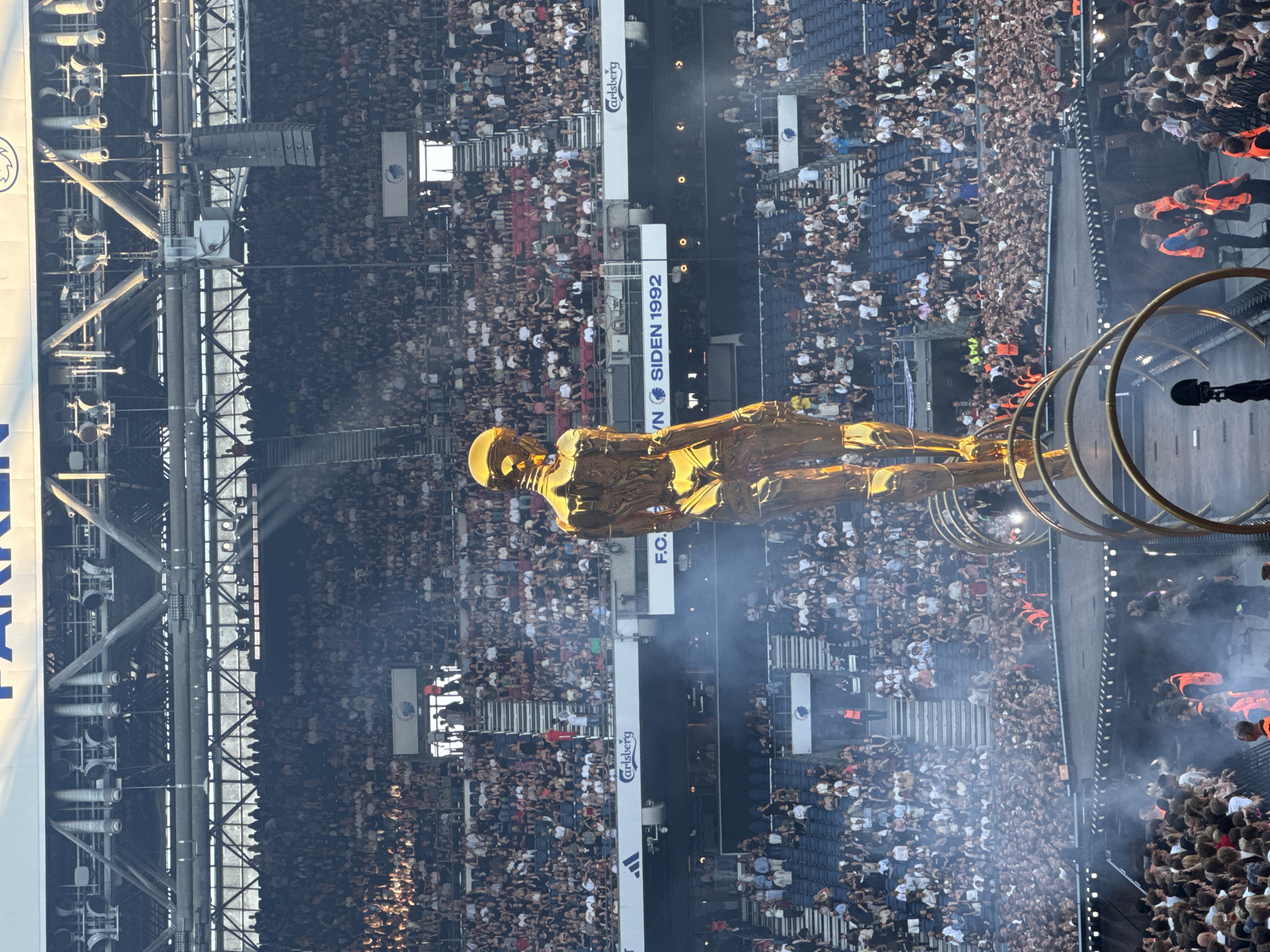 This is a wide shot of a large outdoor stadium filled with a dense crowd of spectators watching an event. In the foreground, a massive, golden, humanoid statue stands center frame, with its back to the camera. The statue is positioned on a stage structure with several large, circular golden elements beneath it. Smoke or fog is visible around the stage. The stadium seating is blue, with a large crowd filling most of the visible sections. Above the crowd, there are banners and structural elements of the stadium, including lighting rigs and large speakers. The text "PARKEN" is visible on a banner at the top of the image, indicating the location is Parken Stadium in Copenhagen, Denmark. The event appears to be a concert or large public gathering.