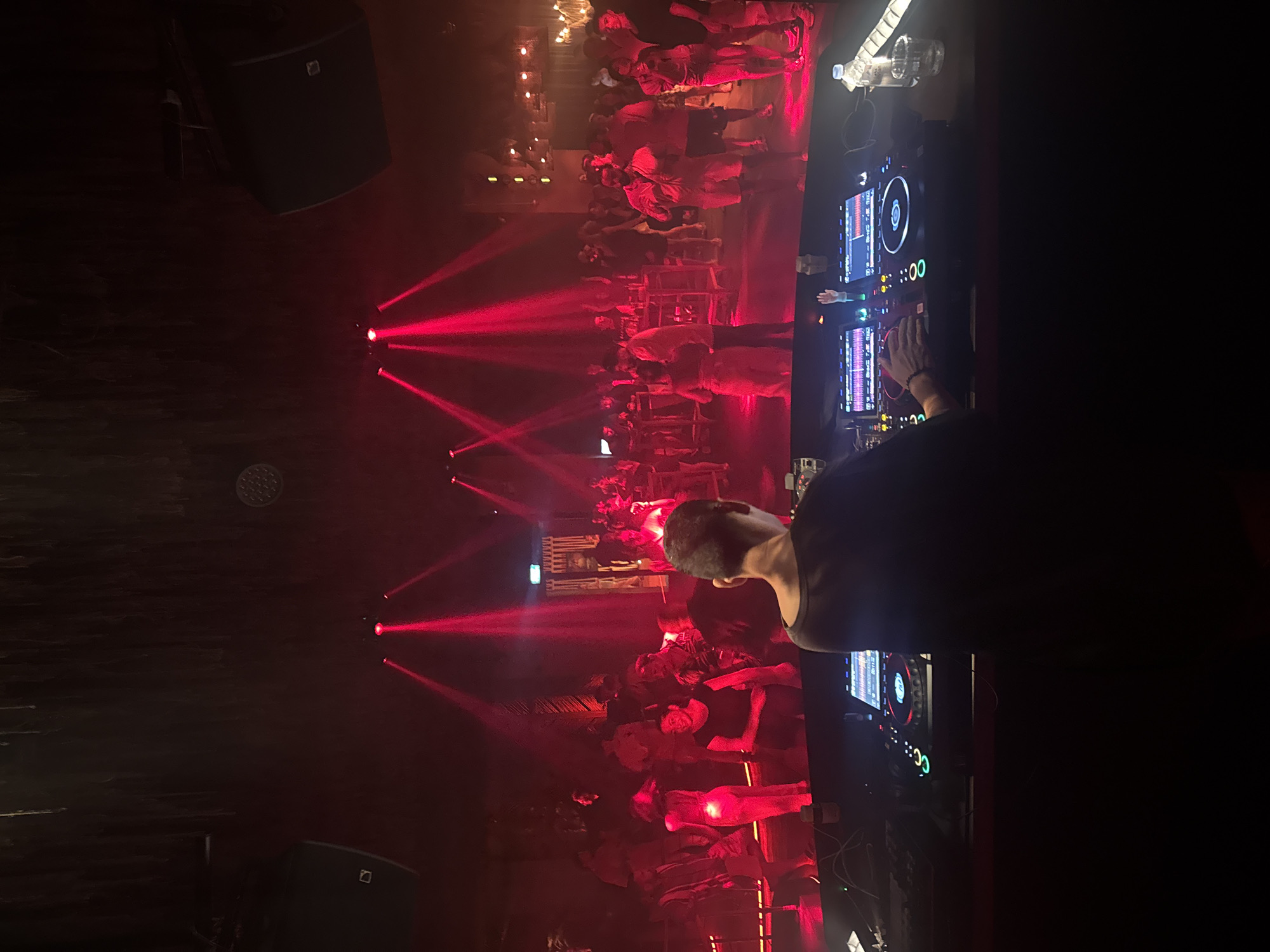 The image depicts a dimly lit nightclub scene in Ban Bang Thao, Thailand, dominated by vibrant red lighting. In the foreground, a DJ stands with their back to the camera, operating a DJ console with illuminated screens and various controls. Their hands are visible on the equipment. Behind the DJ, the nightclub floor is filled with a crowd of people dancing and socializing, illuminated by beams of red light cutting through the haze. Multiple speakers are mounted on the ceiling. The atmosphere appears energetic and lively, characteristic of a nightclub setting.