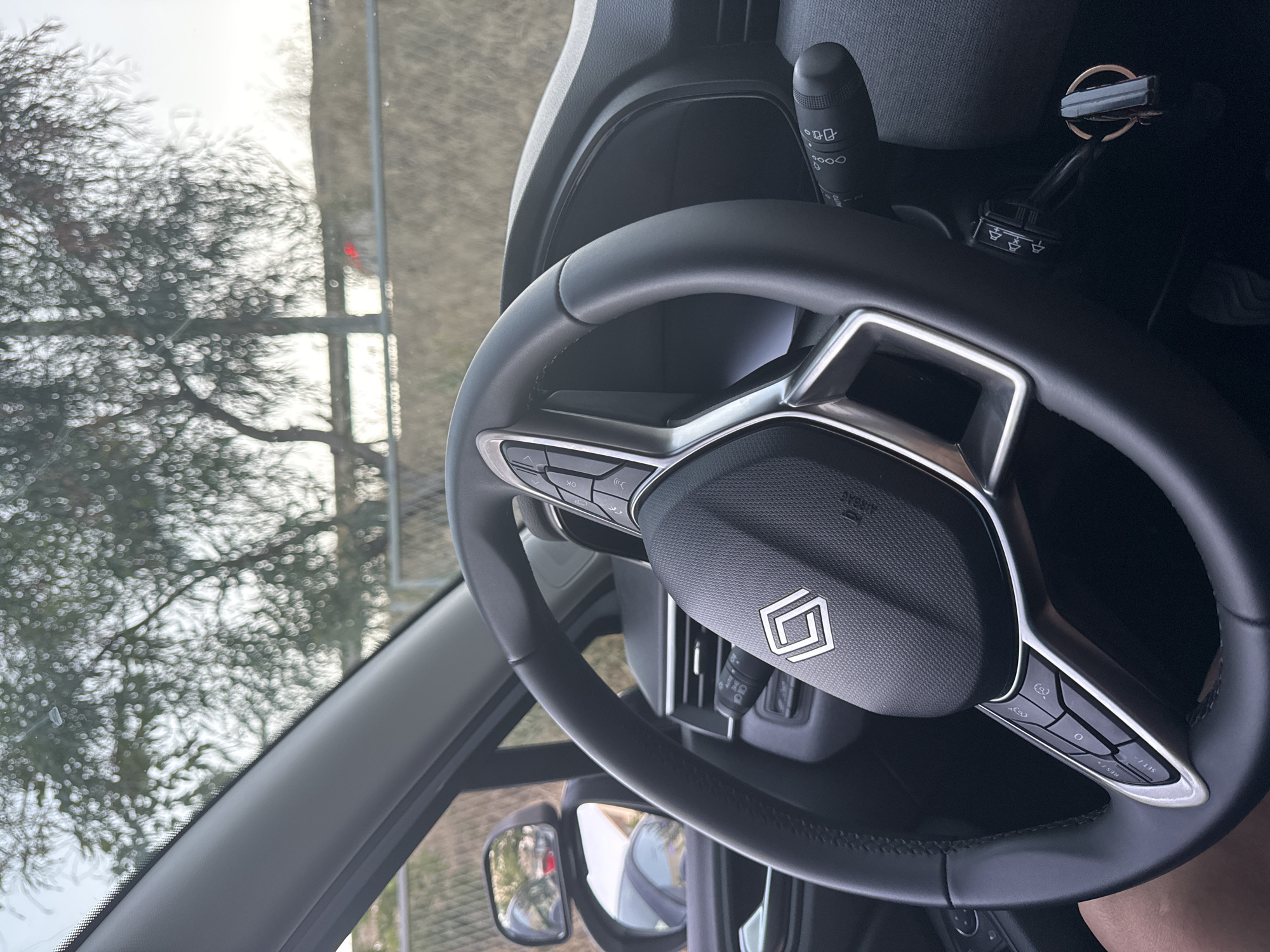 The image shows the interior of a vehicle, focusing on the steering wheel and dashboard. The steering wheel has a distinctive logo in the center, possibly a brand emblem, and various buttons on its spokes. To the right of the steering wheel, a stalk with controls is visible, along with a car key. In the background, through the windshield, a blurry outdoor scene with trees and a fence can be seen. The lighting suggests it might be daytime. The image does not provide any specific visual cues that would allow for the identification of the location within the city of Murcia, Spain.