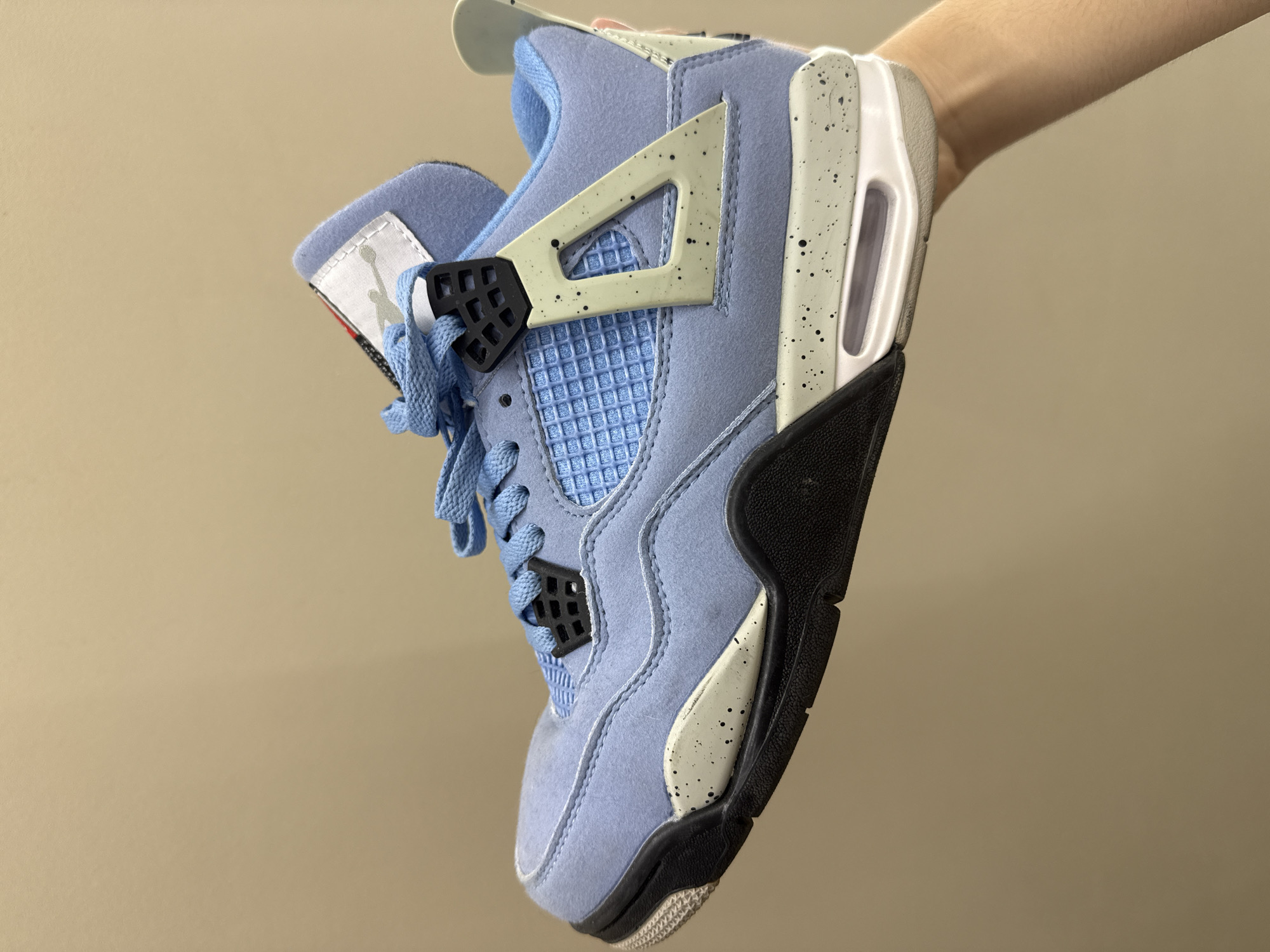 A light blue sneaker with white and black accents is held up by a hand against a plain, tan background. The sneaker is a Nike Air Jordan 4 model. It features a light blue suede upper with a textured blue mesh panel on the side and toe box. Black lace eyelets and a black heel tab are visible. The midsole is white with visible air cushioning and a speckled grey overlay. The outsole is black and grey. The tongue of the shoe has a white tag with a red and grey logo. The background is a solid, neutral color, indicating an indoor setting with no discernible environmental features. The location in Burgas, Bulgaria, is not indicated in the image.