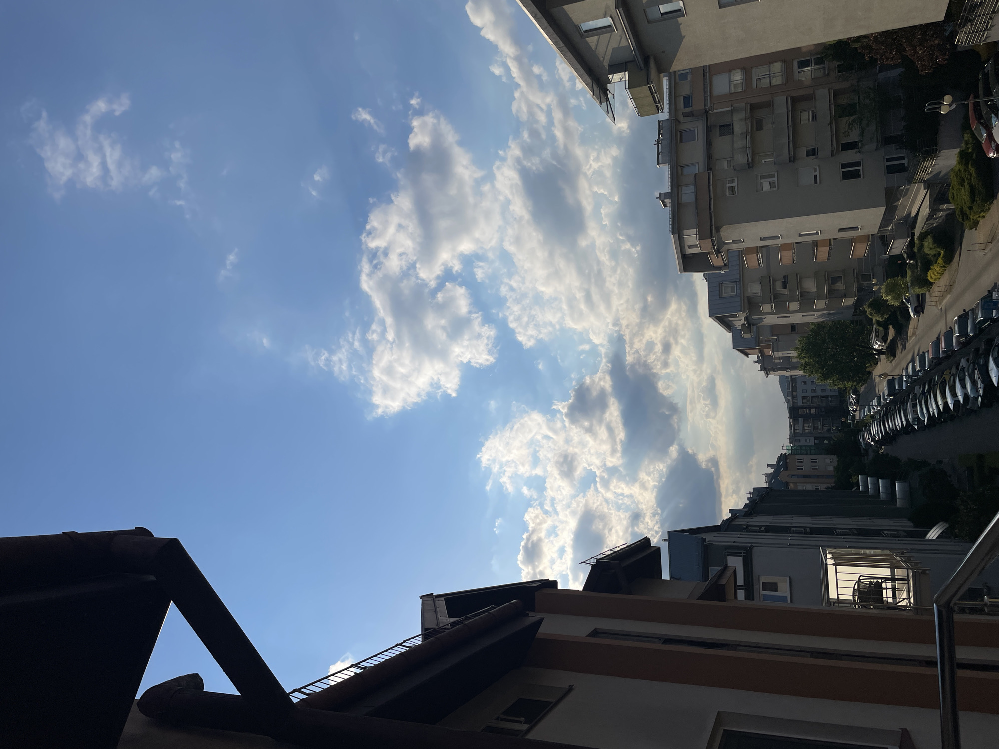 The image shows a view of buildings and the sky in an urban environment. The sky is predominantly blue with scattered cumulus clouds. Sunlight filters through the clouds, creating highlights and shadows. In the foreground are rooftops with visible gutters and architectural details. In the background, taller apartment buildings are visible, lining a street with parked cars. The buildings have multiple windows and balconies. Vegetation, including trees and bushes, is interspersed along the street. The perspective suggests the photo was taken from a lower position, looking upwards. The city appears to be densely populated. The location is likely in Unknown, Unknown, as no specific details are provided in the query.