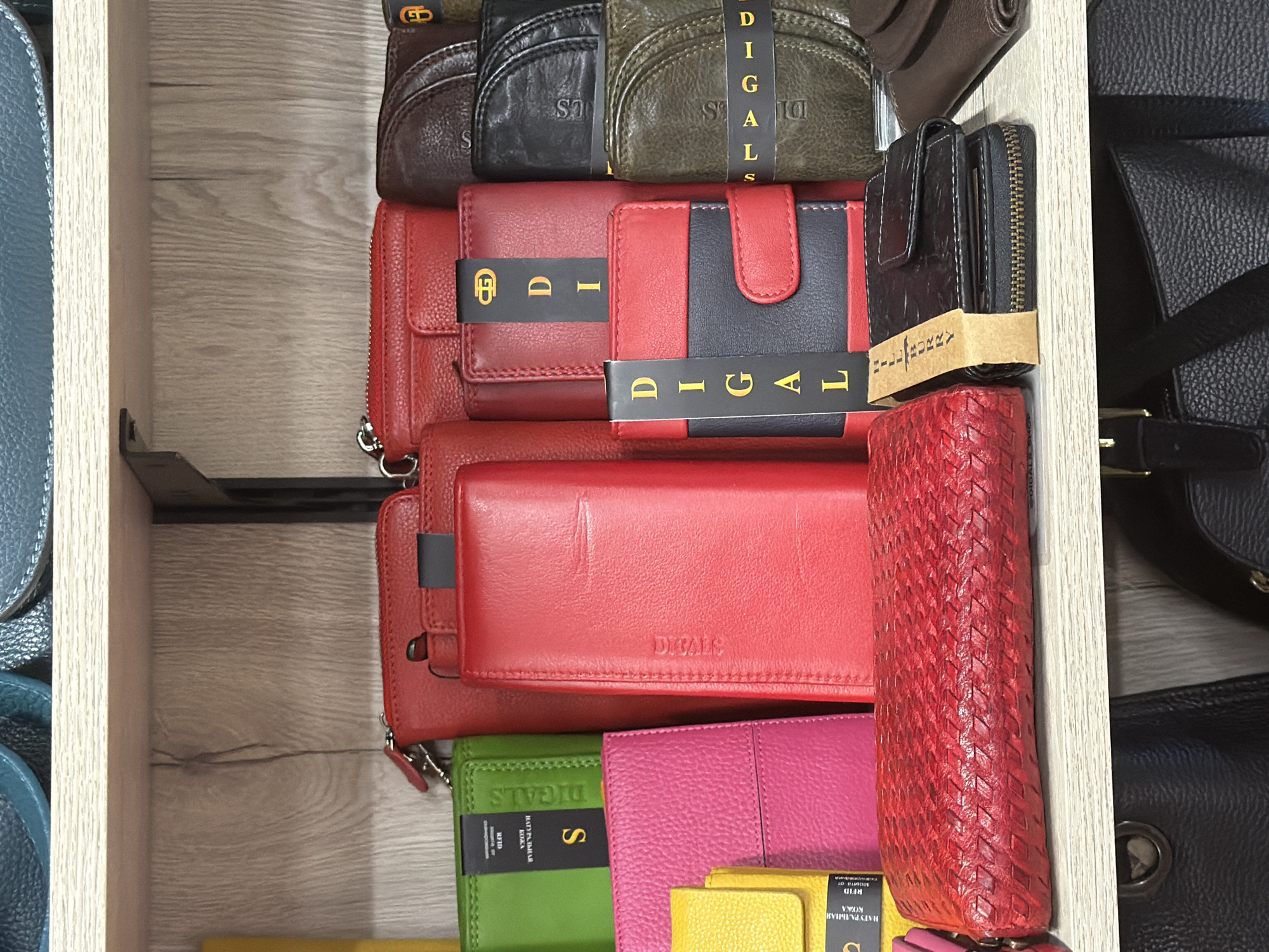 The image shows a close-up, eye-level, interior shot of a retail display shelf filled with numerous leather wallets. The shelves are made of light-colored wood. The wallets are predominantly in shades of red, green, pink, black, and brown, with various textures and designs. Some wallets have visible brand labels or tags. The lighting is bright and even, suggesting an indoor retail environment. There are no people or actions depicted. This scene is consistent with a retail store. While the prompt specifies the city of Sochi, Russia, there is no visual information within the image to confirm this location.