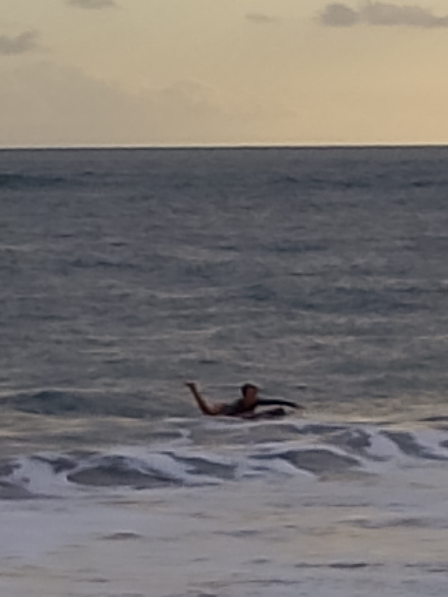 A person is lying prone on a surfboard in the ocean. The person is wearing a dark-colored rash guard and appears to be paddling with their arms. Their legs are extended behind them. The water is choppy with whitecaps. The sky is overcast with a pale yellow hue. The scene is dimly lit, suggesting it is either dawn or dusk.