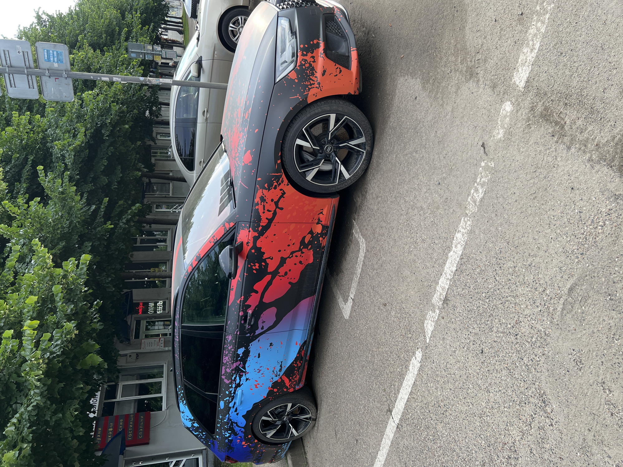 A black Audi A4 Avant station wagon with a custom paint job featuring red and blue splatters is parked on an asphalt surface in Krasnodar, Russia. The car is angled towards the viewer and positioned in a parking space marked with a white line. The car's body is primarily black with a large, abstract design of red, blue, and purple paint splatters extending from the rear of the vehicle to the front fender. The car has black multi-spoke alloy wheels. In the background, a light-colored sedan is parked behind the Audi. Further back, a row of trees with dense green foliage lines the street, and a building with large windows and signage is visible beneath the trees. The overall environment suggests an urban setting on a cloudy day.
