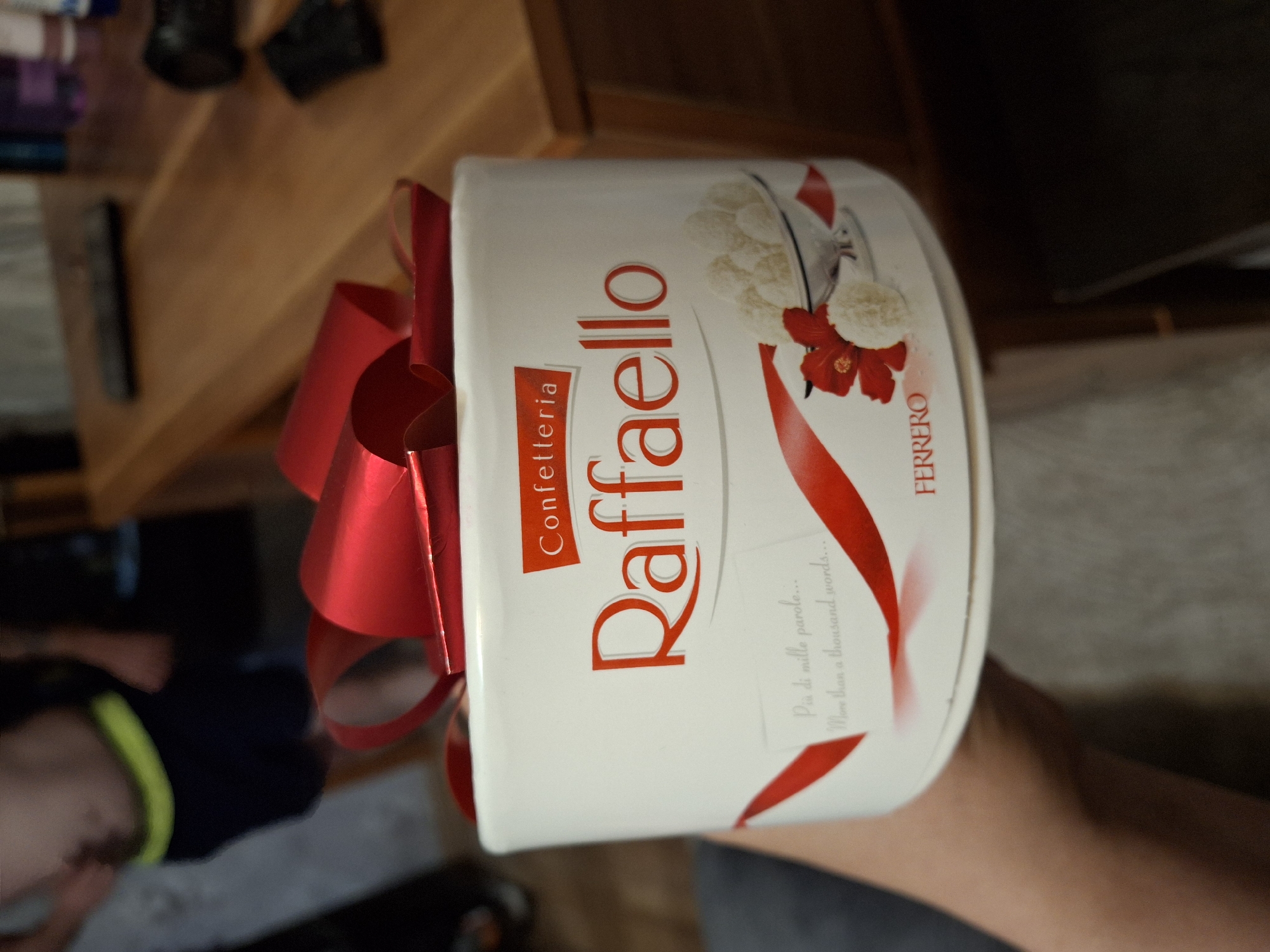 A Raffaello gift box is held by a person in front of a wooden table and a blurred background. The box is white with red lettering that reads "Confezetteria Raffaello" and "FERRERO". It is decorated with a red ribbon and an image of Raffaello candies. The person is wearing dark shorts with a neon yellow waistband. The background appears to be an indoor setting with furniture and some blurry objects. The city of Qarağandı city administration, Kazakhstan is not depicted in this image.