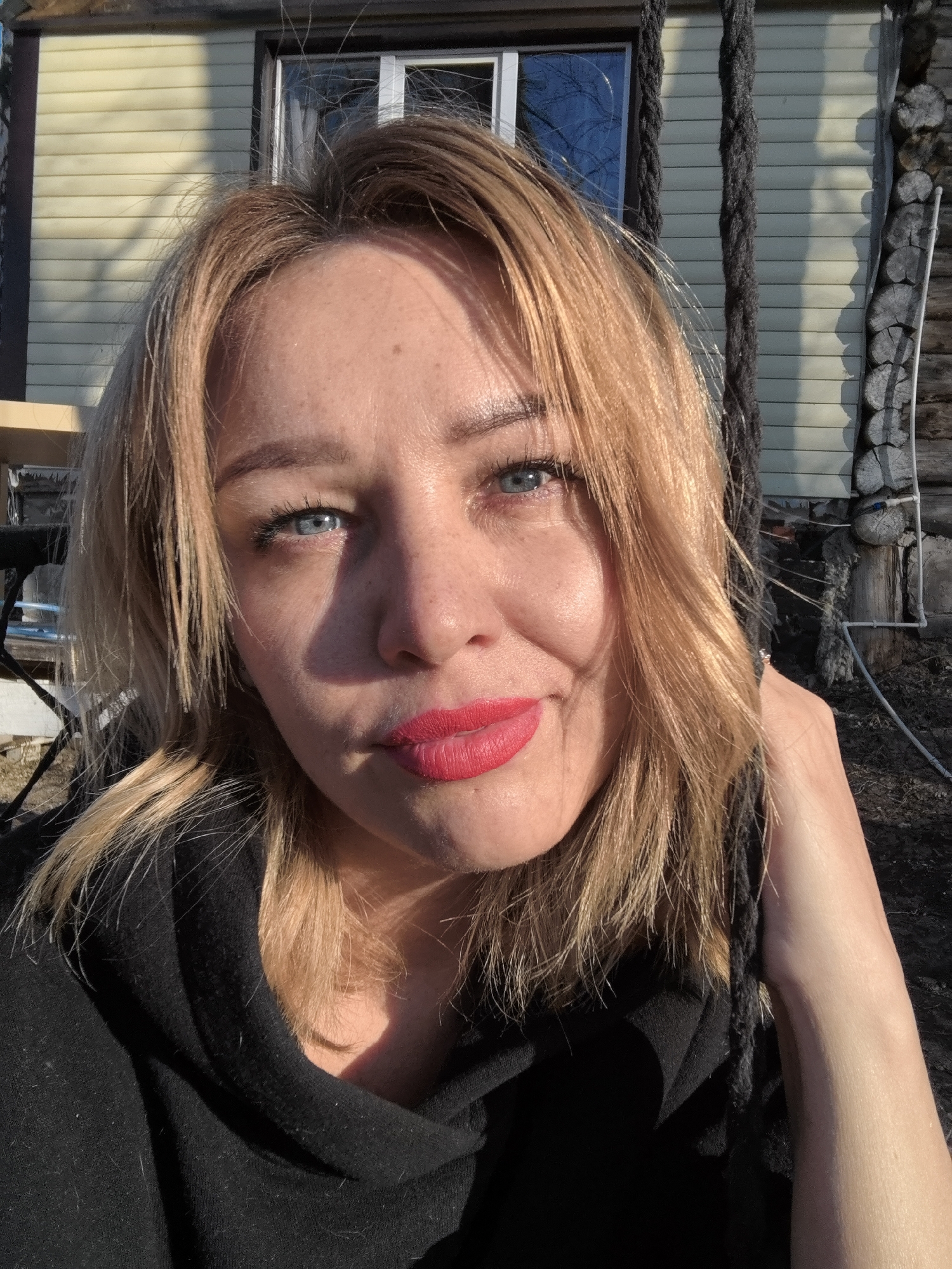 The image is a close-up selfie of a blonde-haired woman with blue eyes and red lipstick. She is wearing a black hooded sweatshirt. The background shows a yellow house with a window reflecting trees. There are also ropes visible on the right side of the image and a wooded area with some snow patches on the ground. The sunlight suggests it is daytime. The location is not discernible to be Noviy, Russia without further information.