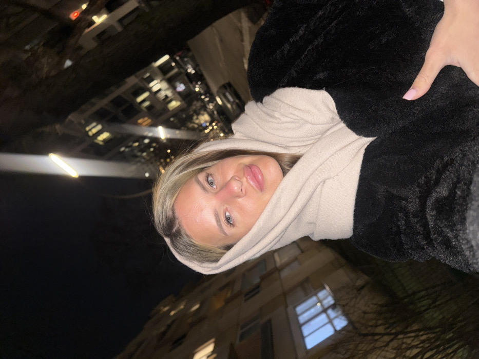 A young woman poses for a selfie at night. She is wearing a black faux fur coat and a cream-colored scarf draped over her head. Buildings and street lights are visible in the background, indicating an urban setting. The image is slightly rotated. The woman's expression is neutral, and she appears to be looking directly at the camera. Her makeup is subtle, emphasizing her lips.