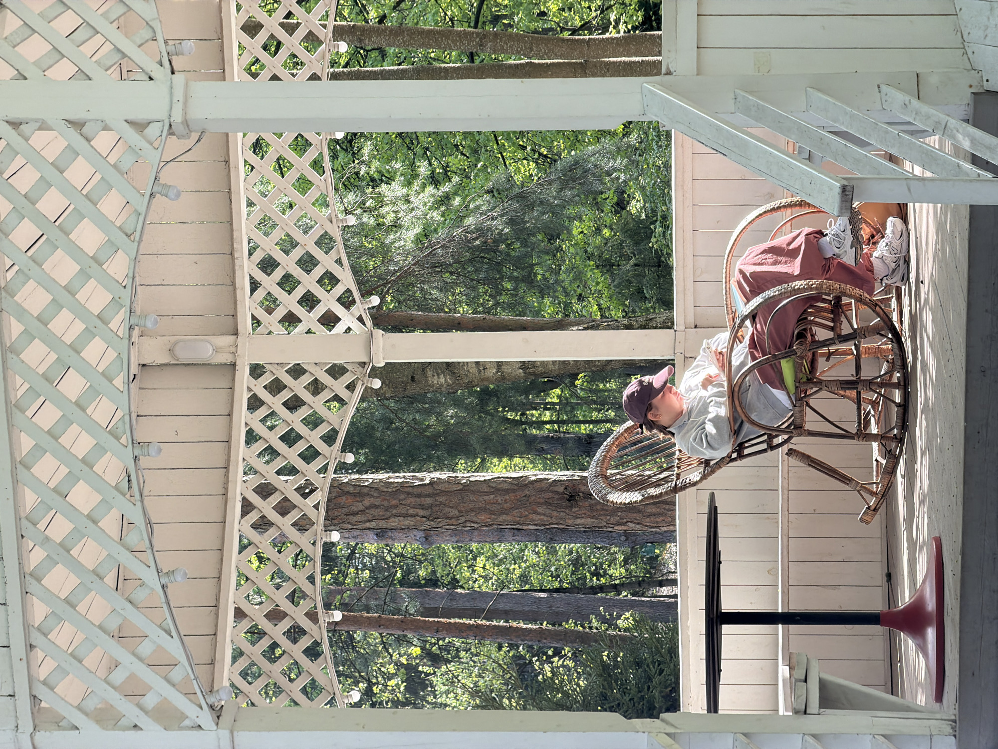 A woman wearing a cap and light-colored clothing is relaxing in a wicker rocking chair on a porch. The porch has white wooden railings and a lattice structure, with light bulbs strung along it. Outside the porch, trees are visible in the background. There is a table with a black stem and a red base near the chair. The location is Vnukovo, Russia.