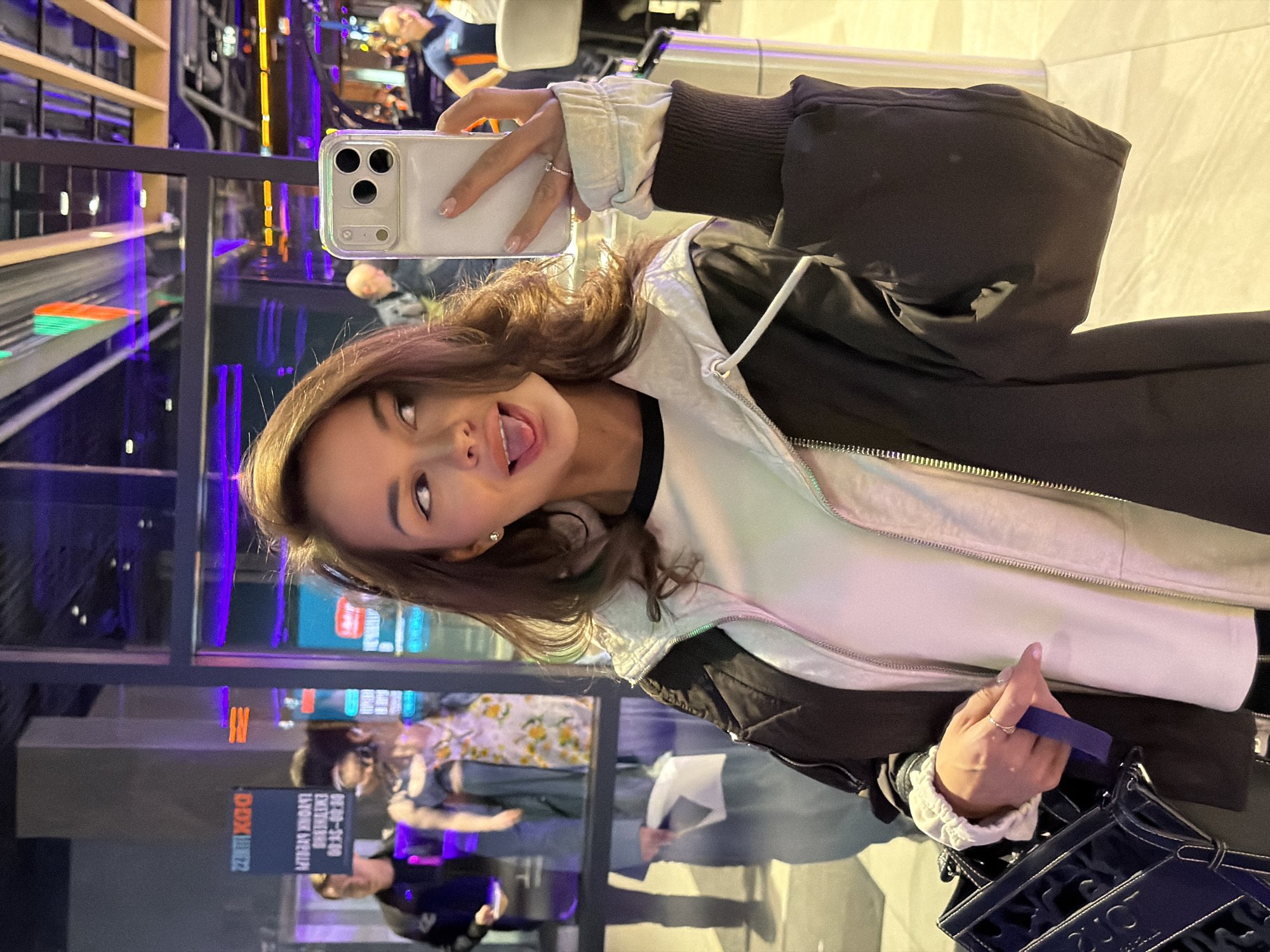 The rotated image captures a young woman taking a playful mirror selfie in an indoor public space in Krasnoyarsk, Russia. She has long, dark hair and is depicted sticking out her tongue and looking upwards with a lighthearted expression. She wears a black jacket over a white inner jacket or top, with visible ruffled cuffs of a light-colored long-sleeve shirt underneath. A silver ring is visible on her left hand, which also features light pink nail polish.
In her right hand, she holds a white smartphone, likely an iPhone, with its triple-lens camera array visible as it faces the reflective surface. She carries a dark blue bag, possibly a tote, with the brand name "DIOR" prominently visible in white lettering.
The background is characterized by numerous reflections in glass or mirrored surfaces, revealing other people and elements of the venue. Bright artificial lighting, including vivid purple and orange neon lights, suggests an entertainment or commercial complex, likely captured in the evening or at night. Reflections show other individuals, including one person in a blue and orange shirt and another wearing a floral patterned top. Visible text on a reflected sign in the background reads "DOX KBB22" (which appears to be reversed) and partially legible phrases such as "08:00-22:00 ENTRANCE ONLY".