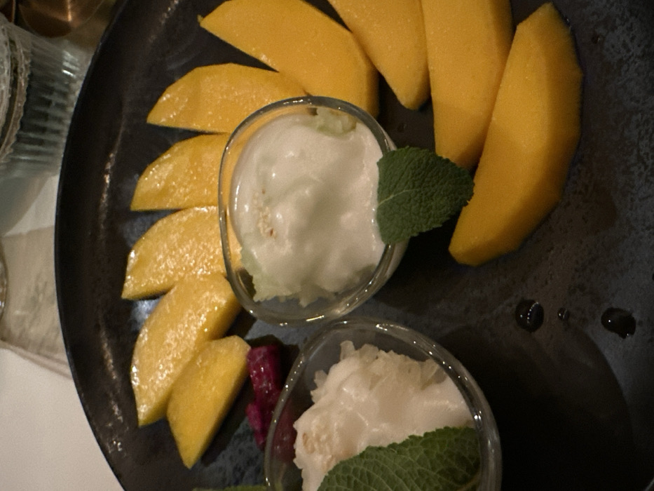A close-up, elevated view captures a dessert served on a dark, possibly matte ceramic, elliptical plate. The plate displays numerous bright yellow, peeled mango slices, arranged in a semi-circular pattern around the right and upper perimeter. Two small, clear glass bowls occupy the center-left. The left bowl contains a white, textured, semi-solid substance, possibly sticky rice, partially adorned with a green mint leaf. The right bowl holds a smooth, white, creamy substance, likely ice cream or mousse, topped with a few small, light brown specks and fronted by another green mint leaf. A small, irregular-shaped, dark pink item, resembling a piece of fruit, rests on the plate between the two bowls and the mango slices. Several small droplets of clear liquid are visible on the dark plate surface. The background is out of focus, showing a light-colored surface and the upper portion of a ribbed clear glass.