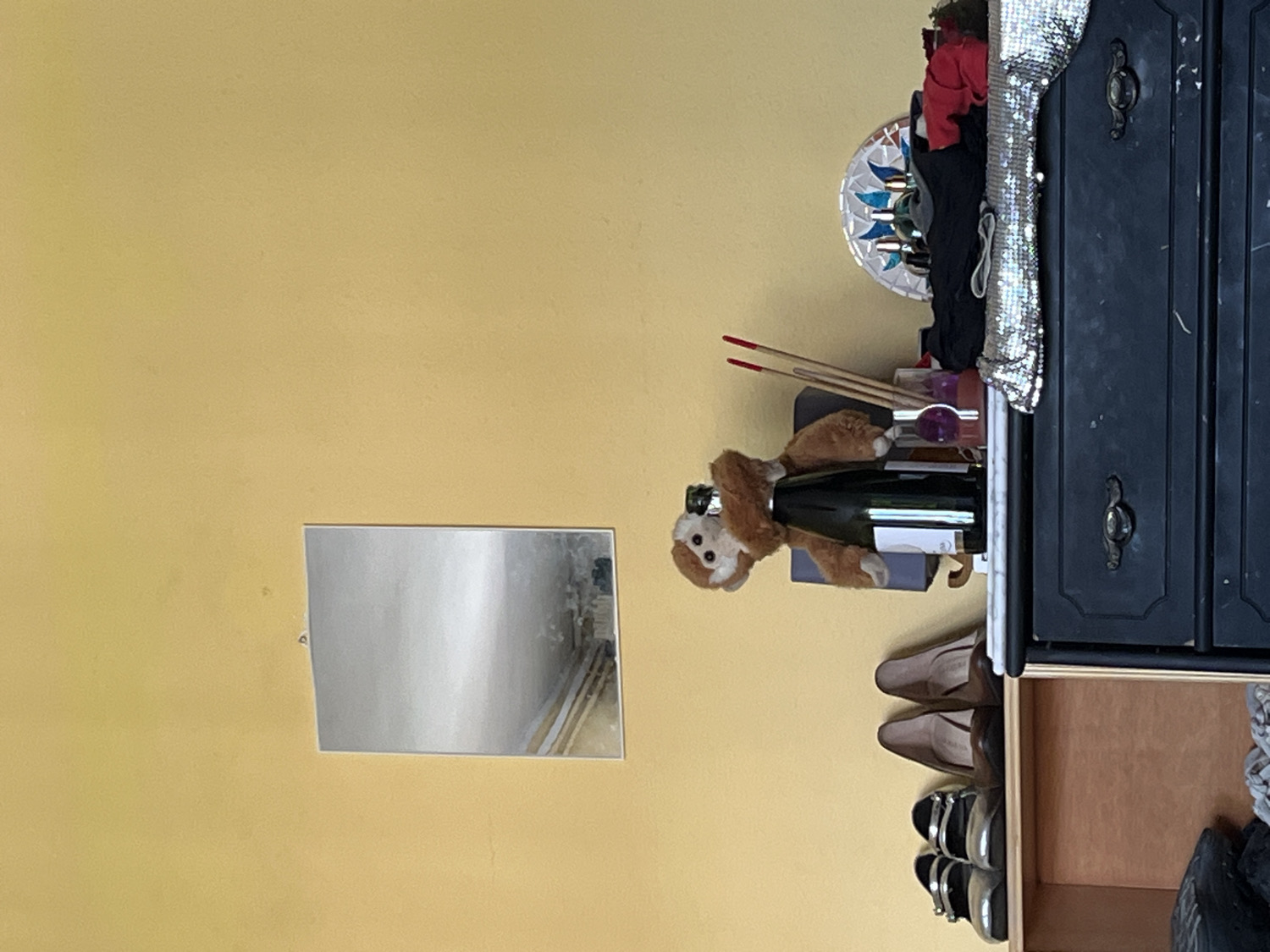 This image captures an indoor scene, likely a personal living space such as a bedroom or dressing area, set against a light yellow-beige wall. The photograph is oriented sideways.
On the left side of the frame, a small, rectangular mirror hangs on the wall. To the right, a dark-colored dresser or cabinet with two dark handles is positioned. The top of the dresser features a decorative arrangement. A dark green bottle, resembling a champagne or wine bottle with a white label, stands prominently, with a small, brown plush monkey toy draped over its neck and top. Beside the bottle, several thin, reddish-tipped sticks, possibly incense, stand upright in a container, alongside other small items including a purple spherical object and a decorative circular plate with blue and white patterns. A shiny, silver, textured fabric item, potentially a handbag or scarf, is draped over the front corner of the dresser.
Below the dresser, an open wooden shelf or drawer reveals several pairs of shoes, including brown heels and silver and black flats. No individuals are present in the image, and no specific activity is occurring; the scene is a static display of personal belongings. The lighting suggests ambient indoor illumination. The white label on the bottle is the only visible text, but it is not decipherable. While the context mentions Barcelona, Spain, no specific visual elements within the image directly indicate this location.