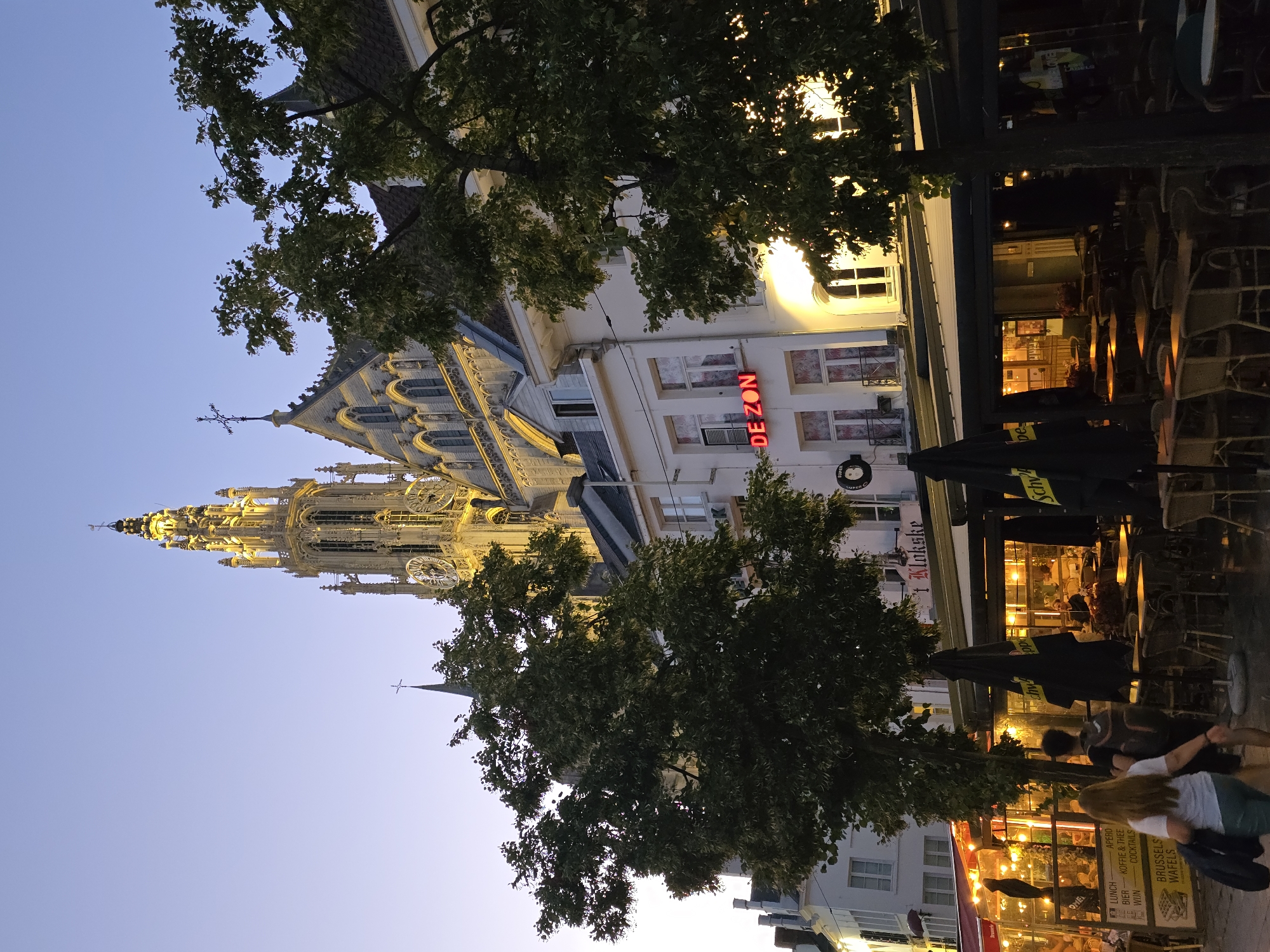 The image depicts a twilight view of a bustling street scene in Antwerp, Belgium, dominated by the imposing spire of the Cathedral of Our Lady. In the foreground, a row of restaurants and cafes line the street, their outdoor seating areas filled with tables and chairs, many under umbrellas. Lights from inside the establishments cast a warm glow. Two people are visible walking on the sidewalk, one with a backpack. Across the street, the facade of a building features a neon sign that reads "DE ZON." The sky above is a gradient of pale blue and purple, indicating the transition from day to night. Lush green trees partially obscure the view of the buildings and the cathedral. The architecture suggests a historic European city.