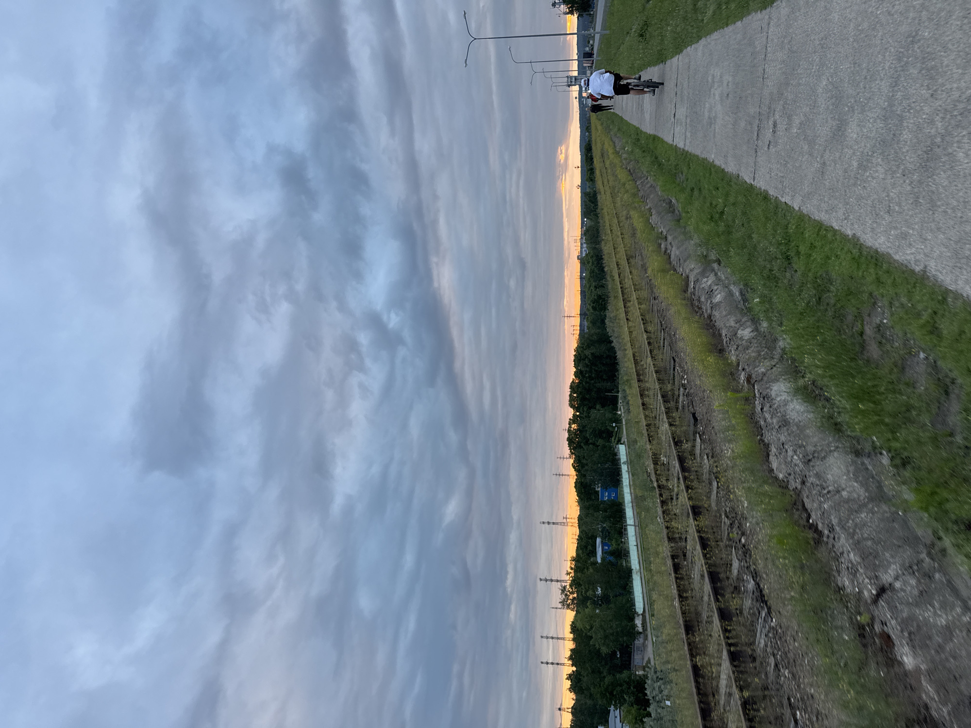 This media file depicts a scenic outdoor view in Chaikovsky, Russia, likely during the late afternoon or early evening due to the soft, diffused light and the warm glow of the sunset on the horizon. The sky is dominated by a dramatic display of clouds, ranging from deep grays to lighter, wispy formations, suggesting a dynamic weather pattern.
The foreground is characterized by a paved pathway running parallel to what appears to be an old railway line or a disused industrial area with rows of weathered structures. Adjacent to the pathway is a strip of lush green grass.
On the pathway, a person is riding a bicycle. The cyclist is wearing a white shirt and dark shorts, and their posture suggests a leisurely ride. Further along the path, more lampposts and possibly power lines are visible, hinting at the proximity of an urban or suburban area. Beyond the railway tracks, a dense line of trees marks the edge of the landscape, with some buildings, including what look like small blue structures, nestled amongst them. The overall atmosphere is serene and tranquil, evoking a sense of peaceful solitude. There is no visible text in the image.