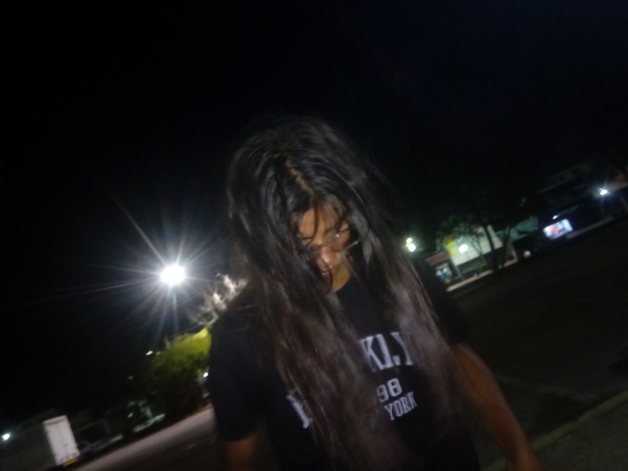 A person with long, dark hair covering their face stands outdoors at night. The person is wearing glasses and a black t-shirt with the text "BKLY 98 YORK" printed on it. There is a light source in the background, possibly a streetlight, causing glare and shadows. The surrounding environment appears to be a street or parking lot with buildings and vegetation in the distance. The location is potentially Juan Griego, Venezuela.