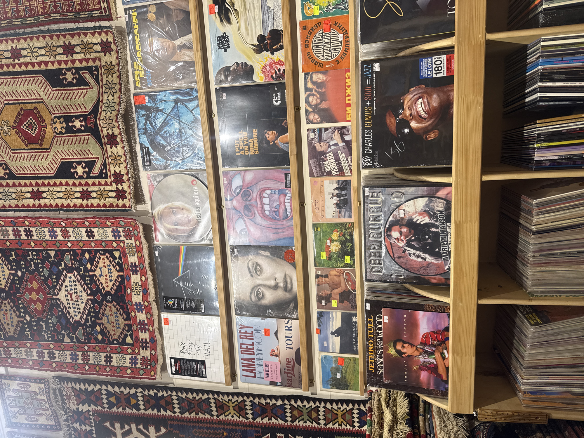 The image shows a record store, likely in Yerevan, Armenia, given the presence of Cyrillic script on some of the album covers. Several vinyl records are displayed in a wooden shelf unit against a backdrop of ornate rugs. The album selection includes a variety of artists and genres, from Pink Floyd's "The Wall" and Lana Del Rey's "Honeymoon" to Nina Simone and Ray Charles. Other visible artists include Deep Purple, Jethro Tull, Marilyn Manson, and Christina Aguilera. The records are stacked both horizontally within the shelves and standing upright in adjacent sections. The rugs displayed on the wall feature traditional geometric designs and appear to be hand-woven. The lighting in the scene is moderately bright.