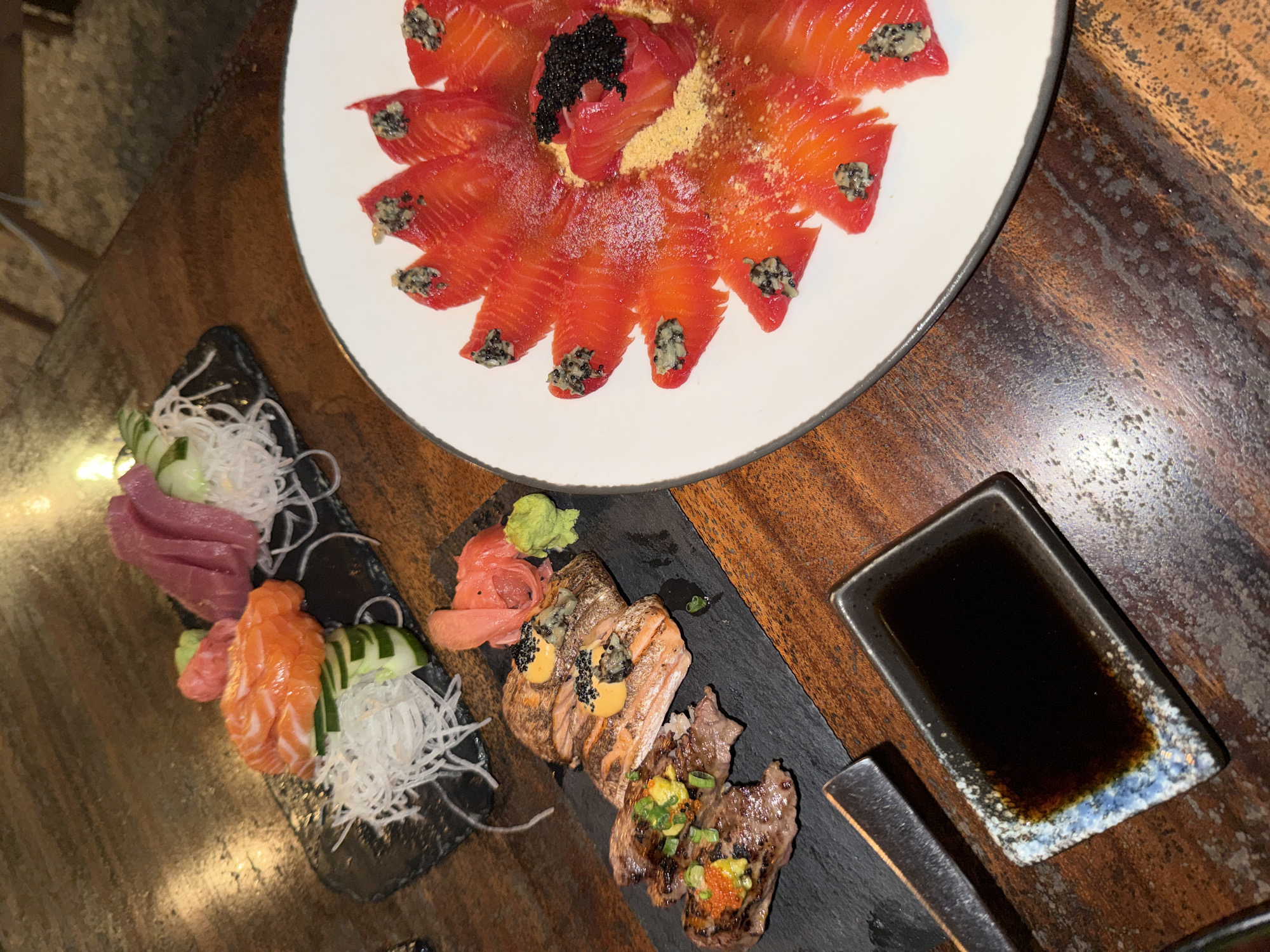 An overhead shot captures a diverse spread of Japanese cuisine arranged on a dark wooden table, indicative of a restaurant setting. The scene features multiple dishes presented on various plates, emphasizing fresh ingredients and meticulous plating.
The prominent dish, centrally located on a large white round plate, showcases thinly sliced, vibrant orange salmon, likely carpaccio or sashimi. These slices are arranged in a circular pattern, forming an edible flower-like design. The center features a mound of salmon adorned with black caviar, while many individual salmon slices are topped with a dark, speckled, almost pesto-like substance. A light, gold-colored powder is dusted across the plate, adding an elegant touch.
To the left, a rectangular black slate plate holds a fresh sashimi selection. It includes several slices of deep red tuna and bright orange salmon, both neatly arranged and garnished with finely shredded white daikon radish and slices of cucumber.
Further right, another black slate plate presents seared items. There are two pieces of seared salmon, possibly aburi sushi or nigiri, each topped with a creamy yellow sauce, small black seeds or roe, and the same dark, speckled topping found on the carpaccio. Beside these are two pieces of seared beef, garnished with finely chopped green spring onions and small orange roe. This platter is accompanied by a dollop of green wasabi and delicate pink pickled ginger.
A small, rectangular, dark ceramic dish containing dark brown soy sauce is positioned on the right. Its rim features a distinctive blue and white speckled pattern. The wooden table surface, with its visible grain and reflections, suggests an indoor environment with ambient lighting. No visible text is present in the image. The setting and dishes are consistent with a restaurant in Kuta Selatan, Indonesia, specializing in Japanese cuisine.