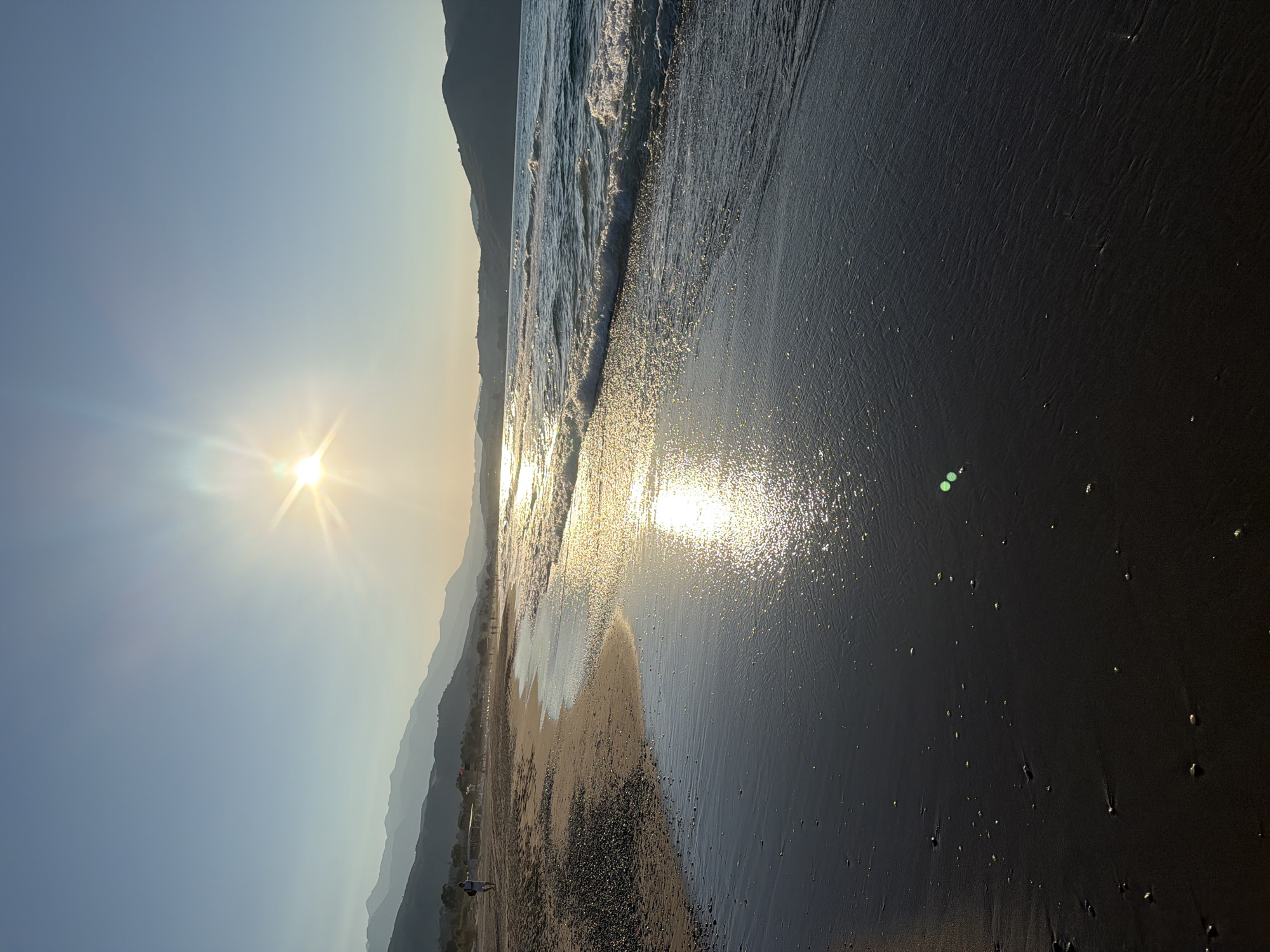 This is a vertical, eye-level, medium shot of a beach in Georgioupoli, Greece, at sunset. The sun is setting behind a distant mountain range and is shining brightly in the upper right quadrant of the frame, creating a lens flare effect and reflecting off the water. The sea is on the right, with waves gently lapping at the shore. The sand is dark and wet in the foreground, with small pebbles scattered on the left side of the beach. There are a few people walking along the shoreline in the distance, and some beach umbrellas and structures are visible further down the beach. The sky is a clear, pale blue. The overall mood of the image is serene and peaceful.