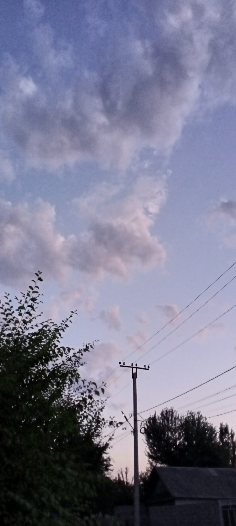 The image depicts an outdoor scene at dusk in Sokuluk, Kyrgyzstan. The sky is a gradient of pale blue and purple, with scattered clouds. A telephone pole with wires stretches across the frame, angled from the lower right towards the upper right. In the lower left, the dark silhouette of a tree with dense foliage is prominent. To the right of the telephone pole, a dark-roofed building with a visible wall is partially obscured by trees. The overall atmosphere suggests a quiet, transitional moment between day and night.
