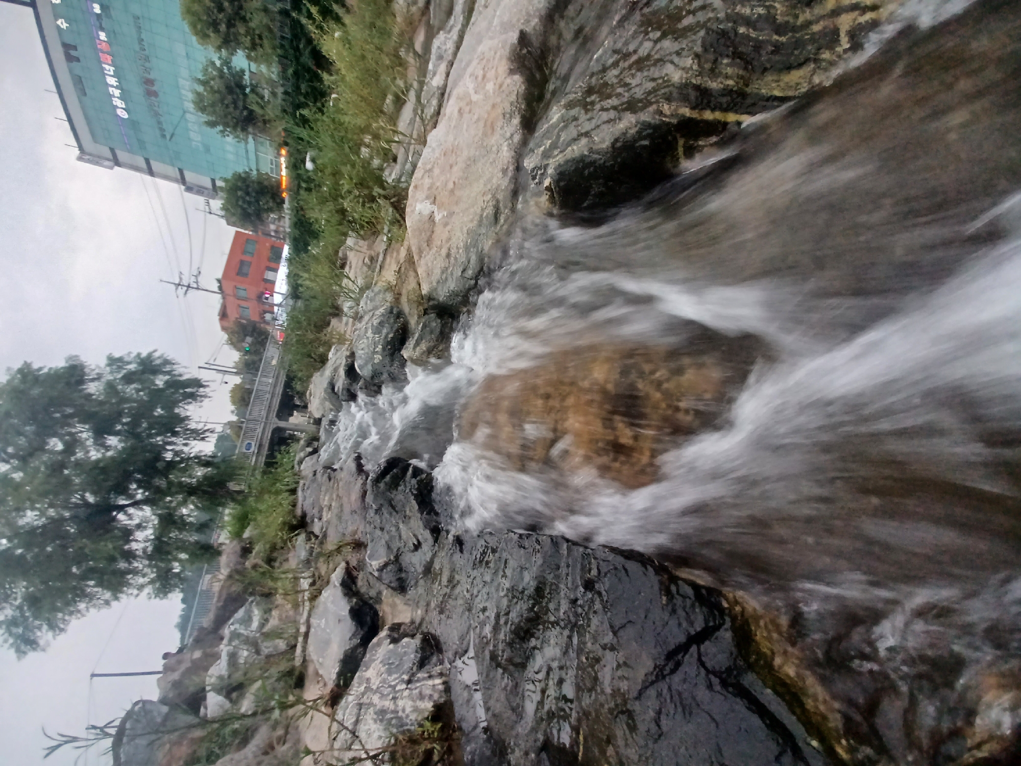 A low-angle, wide shot captures a cascading stream flowing over rocks in the foreground. The water appears blurred due to motion, suggesting a longer exposure. In the background, a bridge spans the stream, with trees and urban buildings visible on the horizon. A large, modern glass building with Korean text and lighting is prominent on the right side of the frame. The sky is overcast, creating a muted lighting condition.