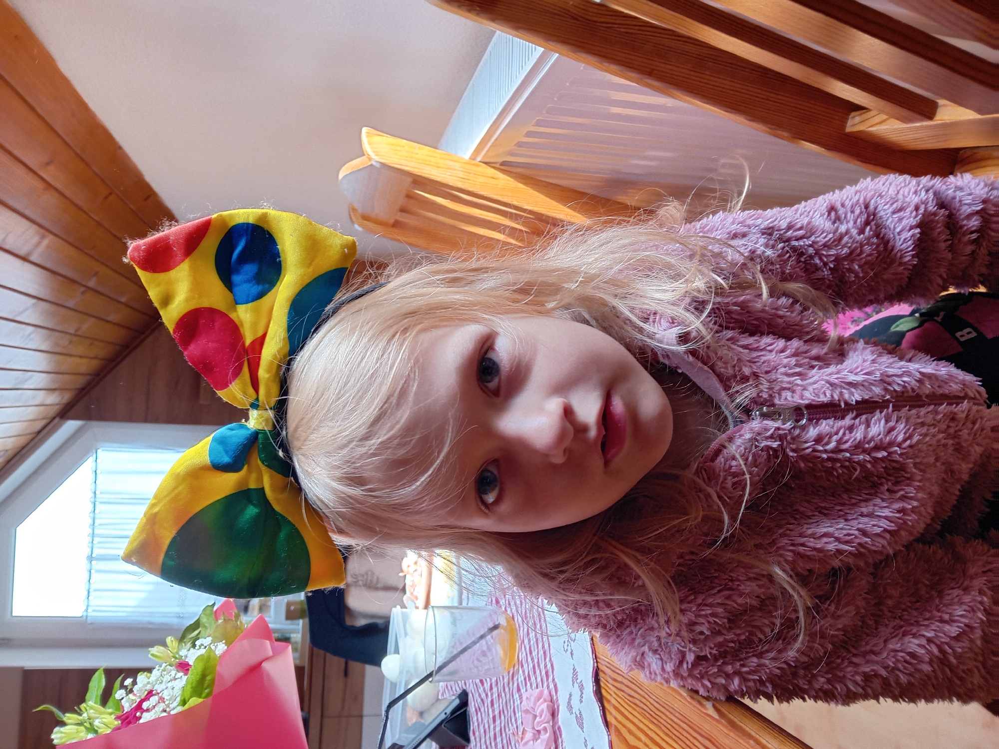 A young blonde girl with blue eyes is seated at a wooden table, looking directly at the camera. She is wearing a fluffy pink zip-up jacket over a dark shirt with a colorful print. On her head, she has a large yellow bow with red, blue, and green polka dots. To her left, a bouquet of flowers is visible, along with a glass of orange juice and a tablet device. The background includes a wooden ceiling, a window with white curtains, a radiator, and wooden chairs. The overall impression is that of a casual indoor setting.
