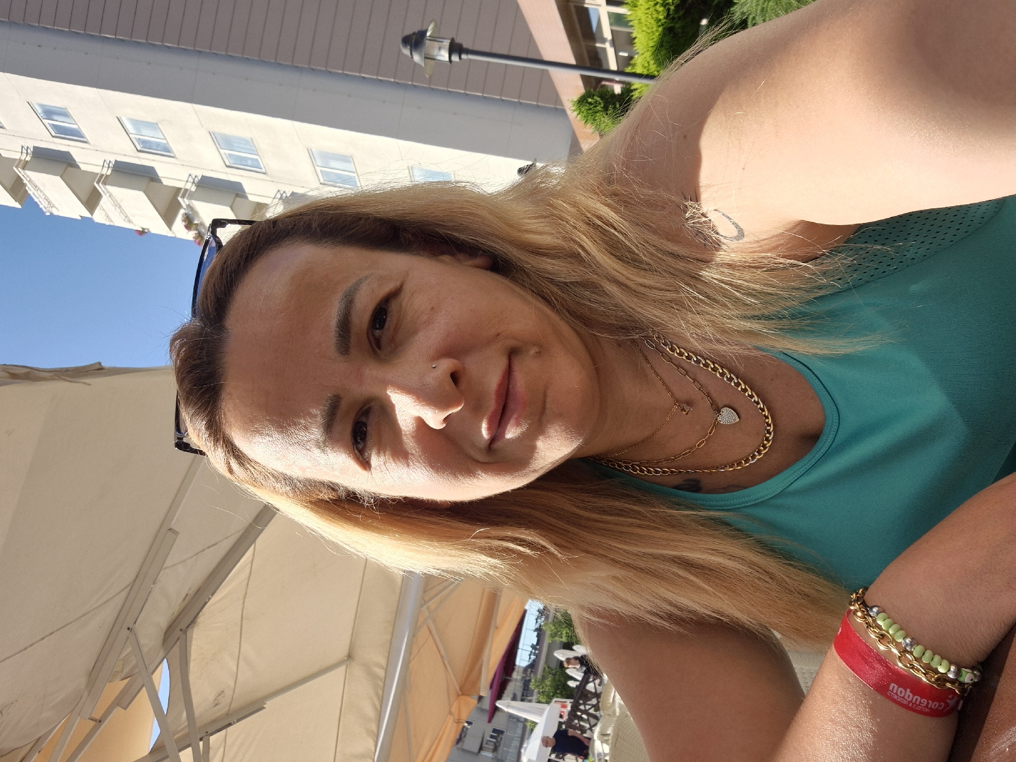 A medium shot captures a woman outdoors, her face turned towards the camera. She has blonde hair and is wearing a turquoise tank top and several necklaces, including one with a heart pendant. Her left arm is visible, adorned with a red wristband and a bracelet. In the background, a tall, modern building with balconies stands against a clear blue sky. To the left, a large umbrella provides shade, and further in the distance, other buildings and people are visible. A street lamp and green bushes are also present in the scene.