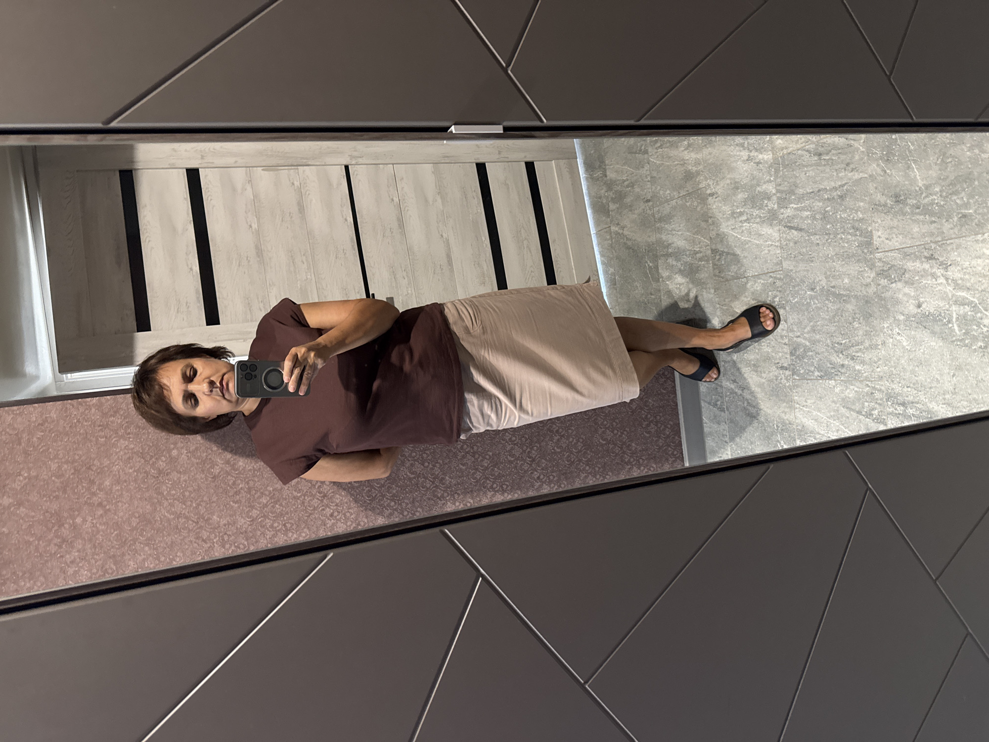 A woman is standing in a room, taking a mirror selfie. She has short brown hair and is wearing a brown t-shirt and a light beige skirt. She is holding a smartphone in her right hand, pointed towards the mirror. The room has patterned wallpaper on one side and a large wardrobe with geometric paneling on the other. A door with horizontal black and white panels is visible in the reflection behind her. The floor is tiled with a grey, marble-like pattern. The image appears to be taken indoors.