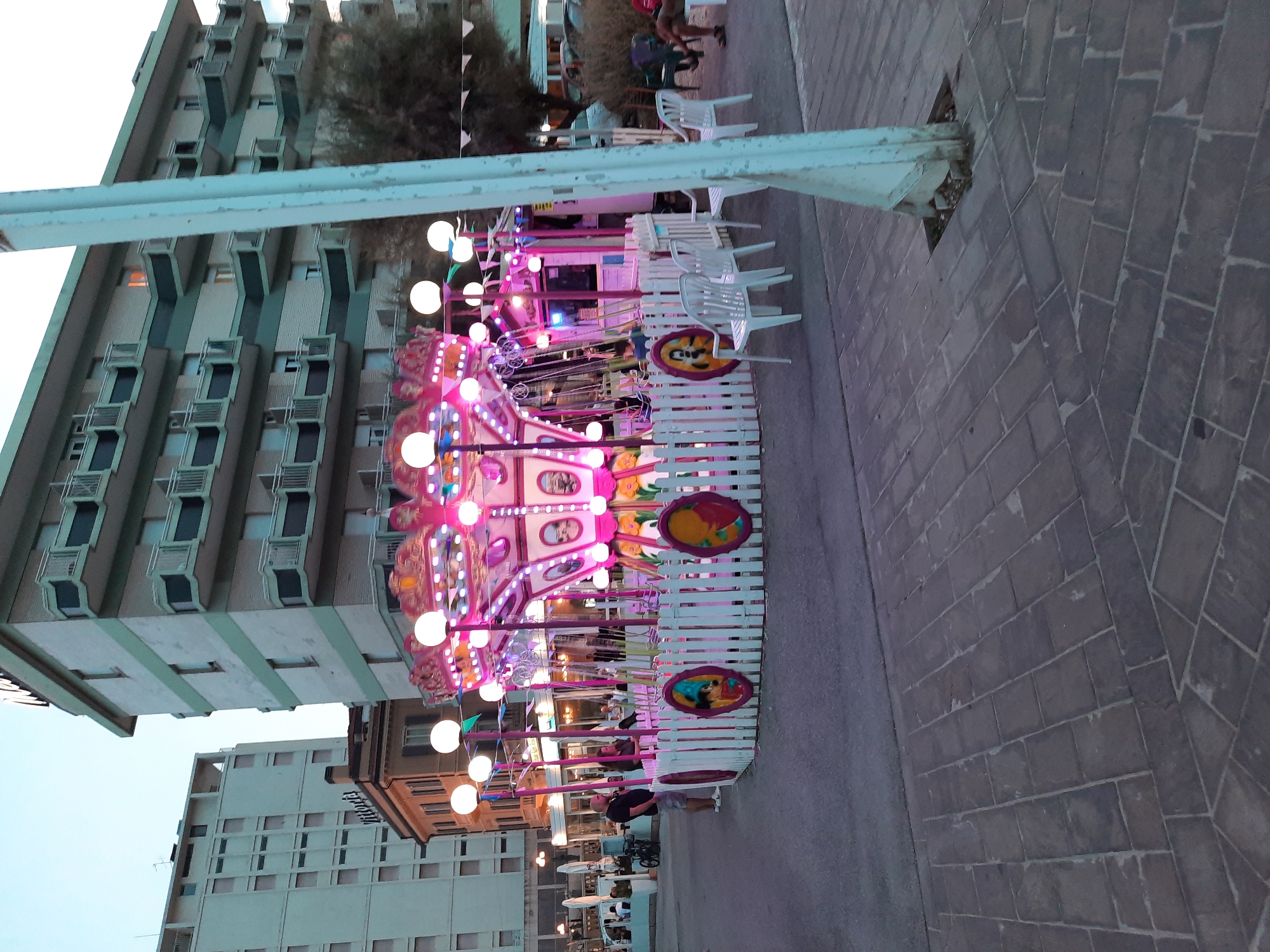 In Pesaro, Italy, on a twilight evening, a small, brightly lit carousel with pink and white accents stands on a paved area. The carousel, adorned with numerous round white lights, features swing-like seats suspended from poles. A low white picket fence with circular decorative panels encloses the carousel. To the left of the carousel, a man in a dark shirt and shorts stands near a bicycle. Further in the background, two tall buildings with balconies are visible. The scene is set against a pale, dusky sky.