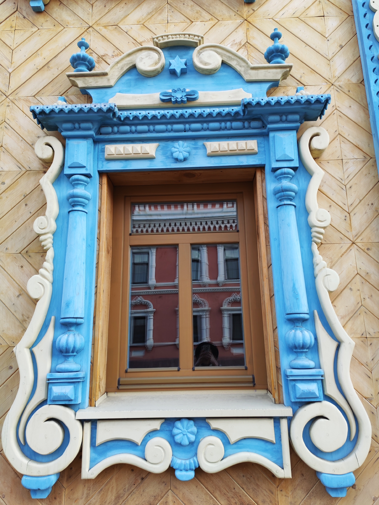 A detailed view of an ornate window in Sarapul, Russia is presented. The window frame is painted a bright blue and accented with white carvings. The frame is adorned with decorative moldings and sculpted floral designs. The surrounding wall is covered in light brown wooden panels arranged in a herringbone pattern. The window reflects an adjacent building with a red exterior, white window frames, and multiple stories. The scene is captured in daylight with a high angle, showing intricate detail and a clear reflection.