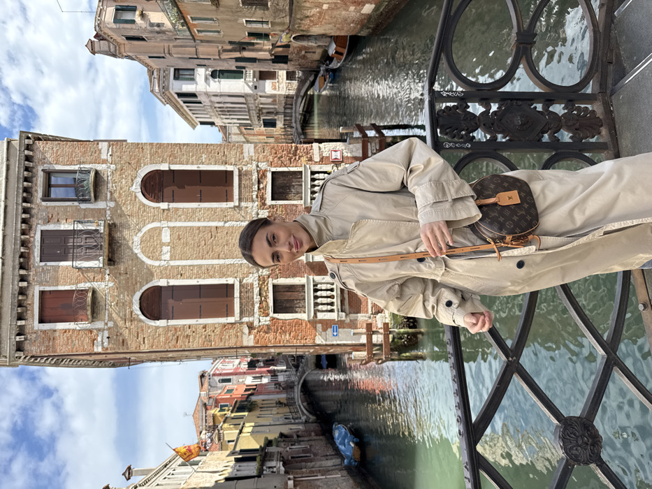 A female individual stands centrally on a bridge in Venice, Italy, facing the camera with a subtle smile. Her dark hair is pulled back, and she wears a light beige trench coat featuring a high collar and shoulder epaulets. A small, round, brown Louis Vuitton monogrammed shoulder bag with a tan leather strap hangs from her left shoulder; her left hand is positioned near the strap, while her right hand is mostly obscured. The bridge's foreground balustrade is an ornate black metal structure with circular decorative elements.
Behind the subject, a canal flows, bordered by multi-story Venetian buildings under a partly cloudy sky. The prominent building directly behind her is a weathered brick structure with multiple windows, closed brown shutters, and two small upper balconies. To the left, the canal extends, revealing additional historic buildings, a small arched pedestrian bridge, and a blue rowboat moored alongside a wooden pole that displays a blue directional arrow sign. A red and yellow Venetian flag, depicting a winged lion, flies from a flagpole on a distant building. To the right, another segment of the canal shows more multi-story buildings and a second arched bridge, with a wooden boat moored. The canal water exhibits a greenish-blue hue.