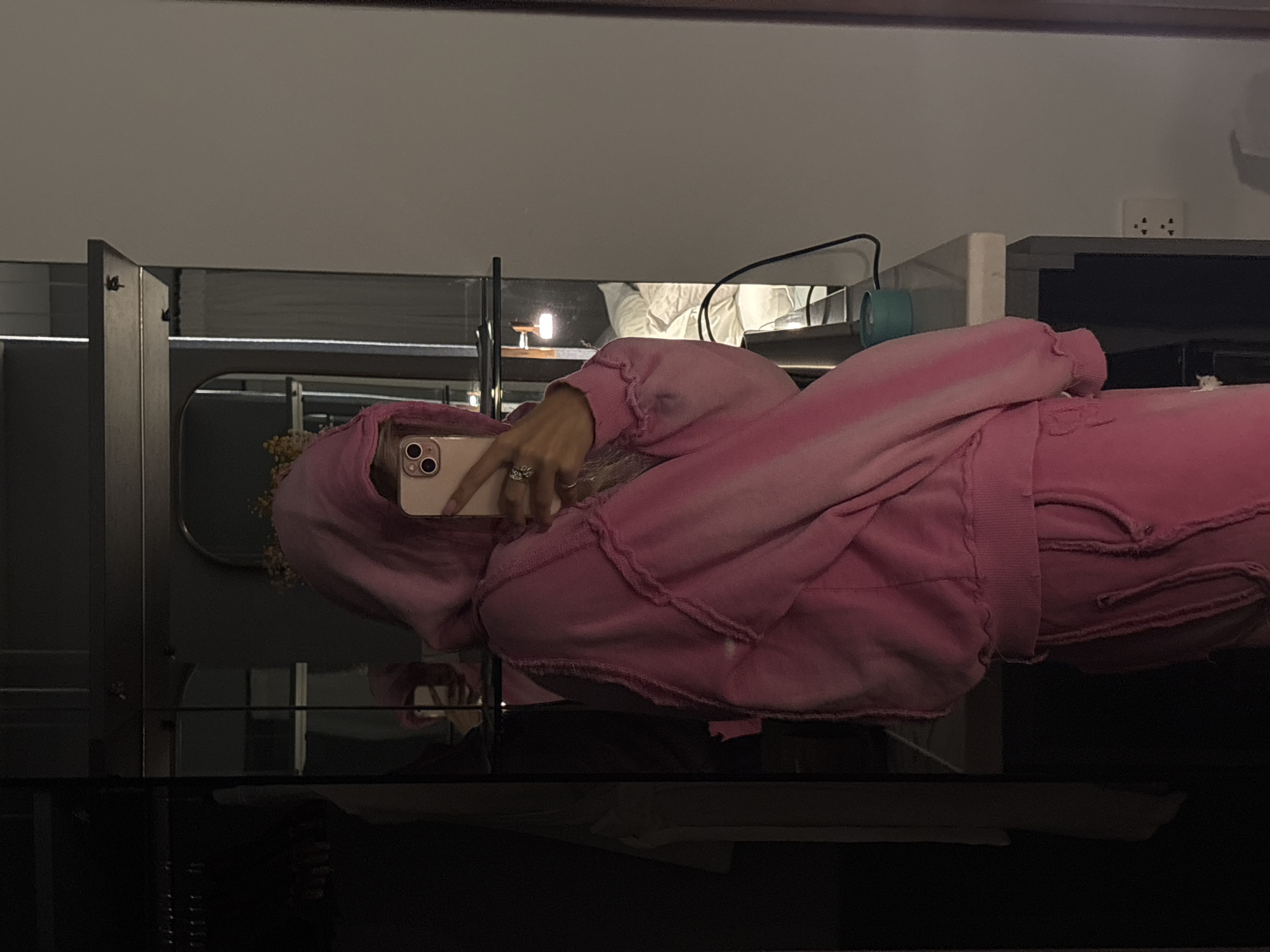 A person wearing a pink tie-dye sweatsuit is taking a mirror selfie in a dimly lit room. The person is holding a pink smartphone in their right hand and is wearing rings on their fingers. The background includes a closet with hangers, a mirror reflecting the person and the room, a shelf with a light fixture, and a bed with white linens. There is also a table with a teal object and electrical outlets on the wall. The overall setting appears to be a hotel room or a bedroom.