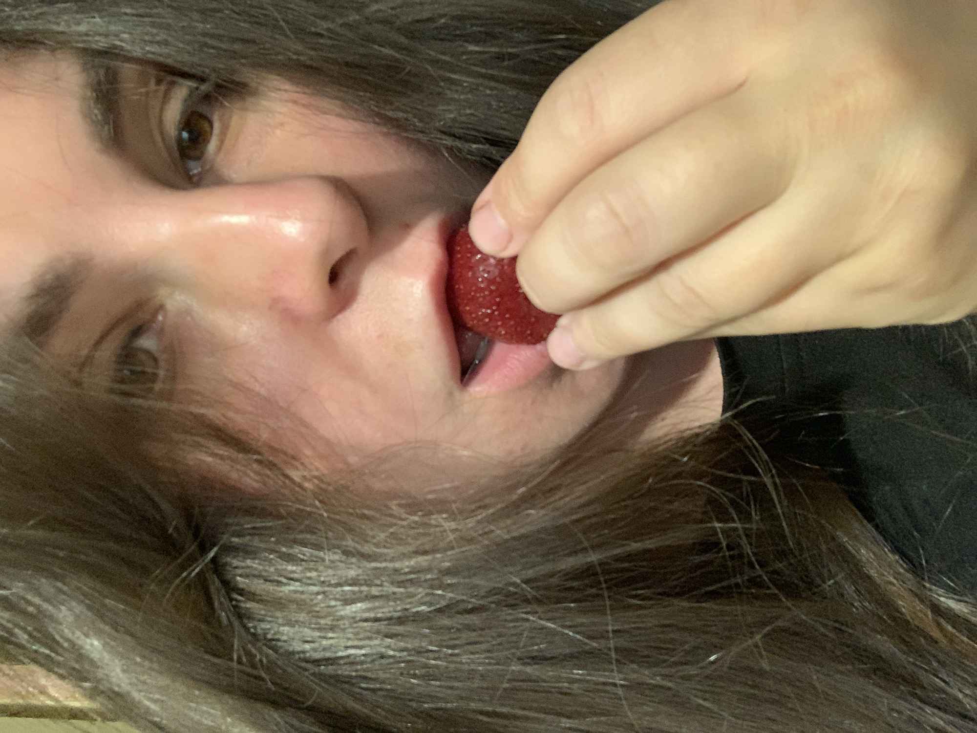 A close-up, high-angle shot shows a person with long, dark brown hair eating a strawberry. The person's face is partially obscured by their hair and the strawberry. Their mouth is open, and their lips are pink. A hand with fair skin and short fingernails is holding the strawberry to their mouth. The person is wearing a black shirt. The background is blurred and appears to be an indoor setting. The scene is dimly lit.