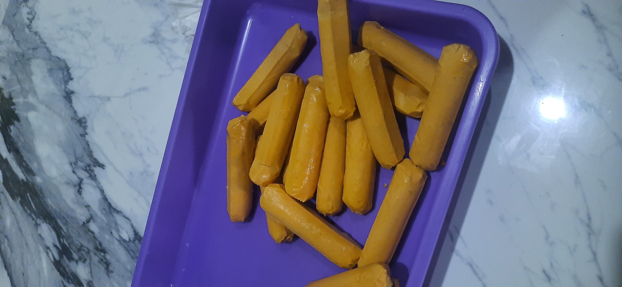 A pile of cooked hot dogs is arranged in a purple plastic tray on a marble countertop. The hot dogs are golden brown and appear to be slightly greasy. The tray is filled with approximately 15-20 hot dogs, some overlapping others. The background shows a close-up of the marble countertop, with dark gray veining on a white background. The lighting is overhead, casting some shadows on the hot dogs. The scene is indoors, likely a kitchen or food preparation area. It is not possible to determine the specific location within Cisauk, Indonesia based on the provided image.