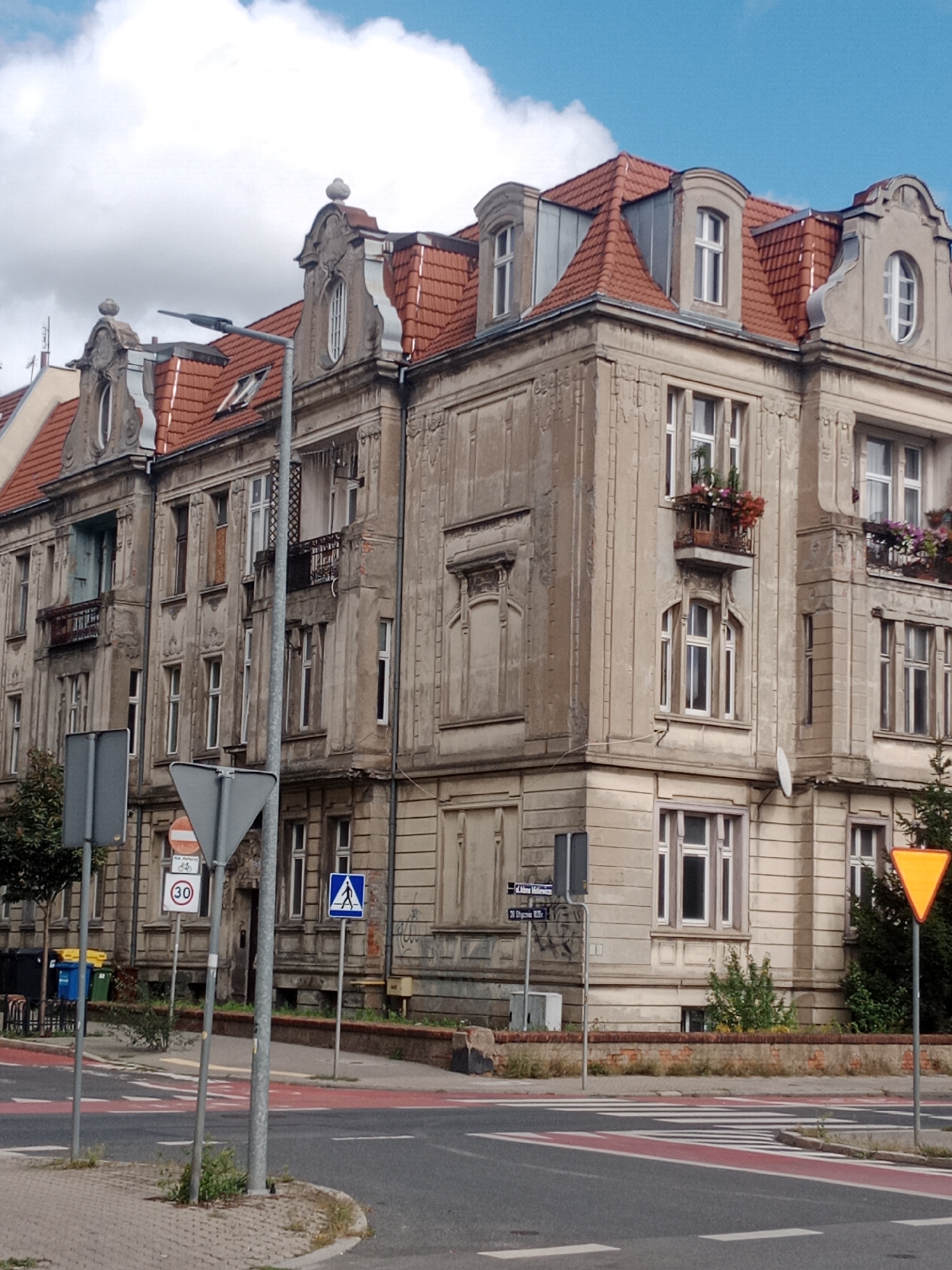 The image depicts a street scene in the city of Bydgoszcz, Poland, focusing on an older, ornate building with a red-tiled mansard roof. The building exhibits detailed architectural elements, including decorative window frames and balconies with wrought-iron railings. Several windows are visible, some with flowering plants on the balconies. The facade is weathered, suggesting its age.
In the foreground, a street with a pedestrian crossing marked by red and white stripes runs across the image. Several street signs are present, including a circular sign with "30" indicating a speed limit, a triangular yield sign, a blue square sign with a pedestrian symbol, and two street name signs. A lamppost stands near the center of the image, and a traffic light pole is visible to the left. Green and blue trash bins are also situated near the building on the left side. The sky above is partly cloudy, with blue sky visible between white clouds. The overall impression is a typical urban streetscape with historical architecture and functional traffic infrastructure.