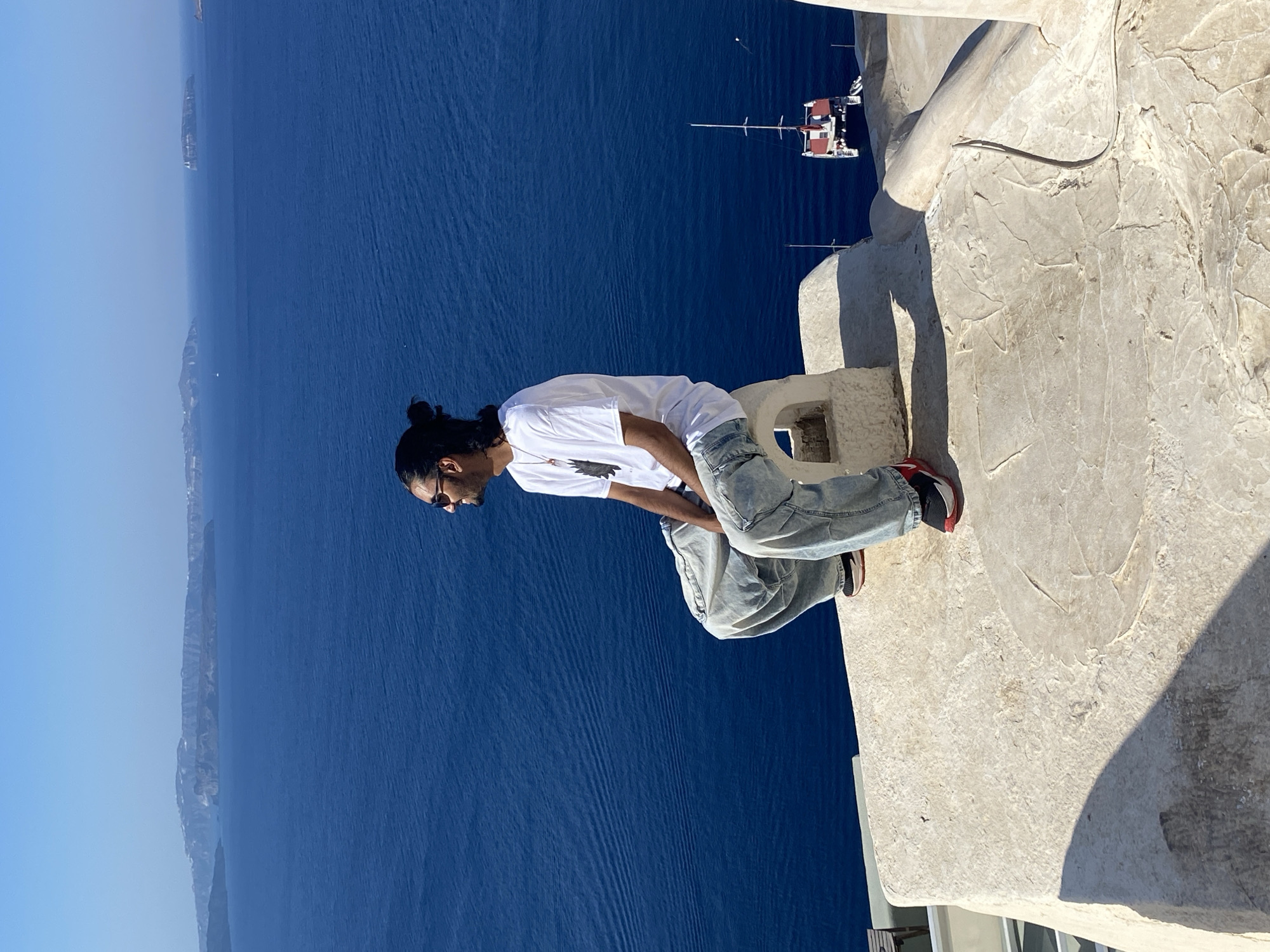 A young man with dark hair pulled into a bun and sunglasses sits on a white stone ledge overlooking the Aegean Sea from Ia Municipal Unit, Greece. He is wearing a white t-shirt and light-wash baggy jeans. His feet are clad in red and black sneakers. To the right of the man, a white catamaran with a red accent sails on the blue water, with landmasses visible in the distance. The sky is clear and blue. The sun casts a shadow from the man onto the textured stone ledge.