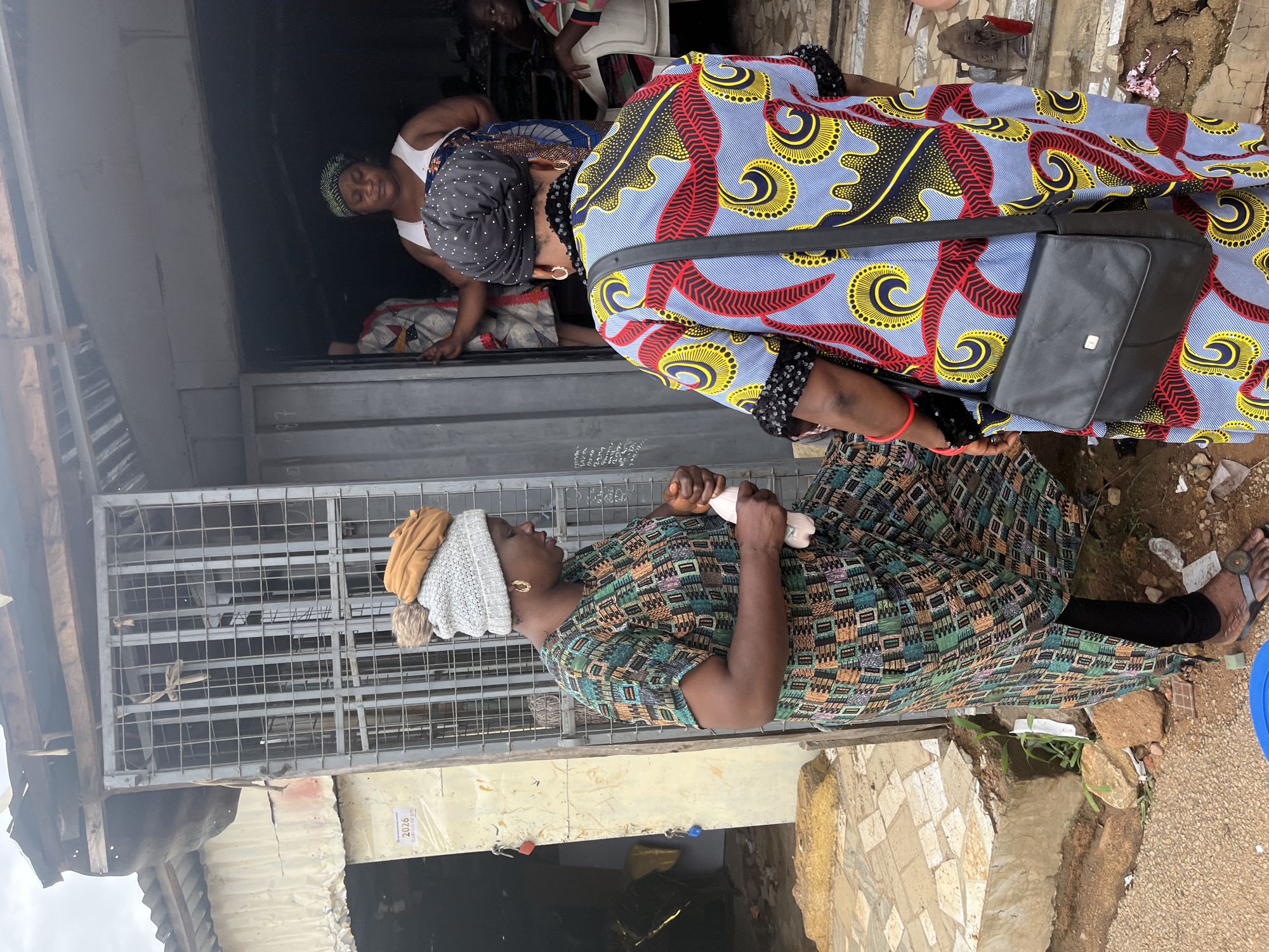 The scene depicts a street in Jos, Nigeria. An African woman, clad in a green patterned dress and a beige and gray headwrap, stands on a stone platform outside of a shop secured by a metal gate. She is holding a white object. Behind her and slightly above stands another woman in a vividly patterned blue, red, and yellow dress with a black bag. A third woman wearing a white tank top and head covering is visible inside the shop. The environment is urban, with the shop being a part of a building showing signs of wear and tear. There is visible waste on the ground near the platform.