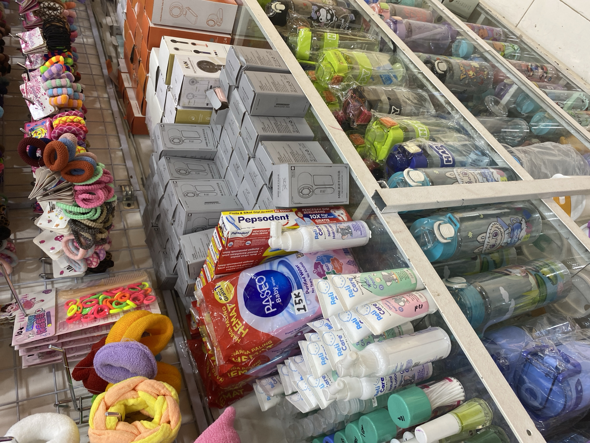 The image depicts a retail shelf displaying various merchandise, likely in a store in Lamteumen Barat, Indonesia. The top shelf features an assortment of colorful hair accessories, including scrunchies and hair ties in a variety of bright hues like pink, yellow, orange, purple, and green. Below this, a selection of boxed items, some with images of what appear to be handheld fans, are neatly arranged. The middle shelf is dominated by baby care products such as wet wipes, lotions, and creams, with prominent branding like "Paseo Baby Wipes" and "Reli Care." Adjacent to these are packaged toothpastes. The bottom shelf is filled with an array of water bottles in different sizes, colors, and designs, some featuring cartoon characters. The overall environment suggests a well-stocked shop offering a diverse range of consumer goods. There are no people visible in the image.