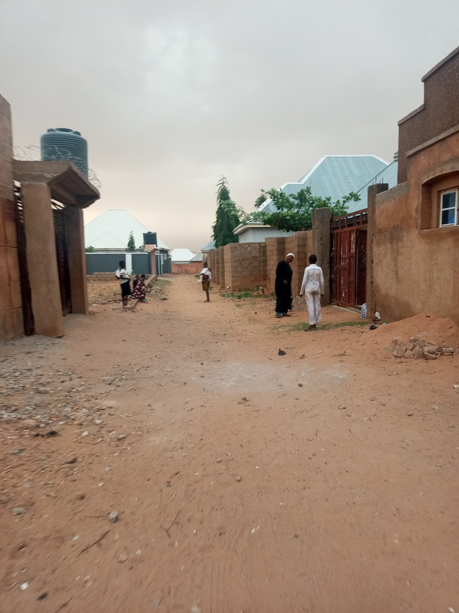 The image depicts a dusty street in an unidentified neighborhood, under an overcast sky. The street is unpaved and composed primarily of packed reddish-brown soil with scattered debris. Buildings with earth-toned facades line the sides of the street, some with gates. A large water tank is visible on the roof of one building. Several people are present. On the left side of the street, three children are standing or sitting. Further down the street, two adults are interacting. One is wearing a black robe, and the other is wearing a white outfit. A water bottle is visible near a gate. The overall environment appears somewhat impoverished.
