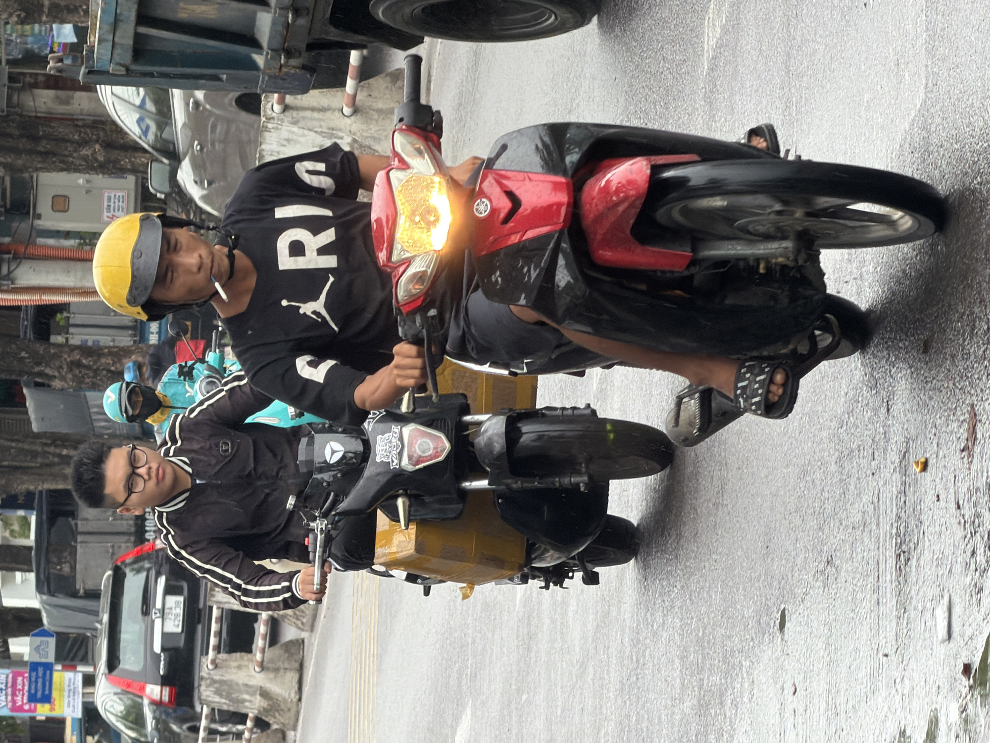 The image captures a street scene in Nha Trang, Vietnam, on a wet day. In the foreground, two men are riding motorbikes. The man on the right, wearing a yellow helmet and a black t-shirt with white lettering, is smoking a cigarette and riding a red and black motorbike. The man on the left, wearing glasses and a black jacket with white stripes, is riding a black motorbike with a large yellow box strapped to the back. Both motorbikes have their headlights on. The road is wet, indicating recent rain, and there are concrete barriers and trees visible in the background, along with other vehicles and buildings.