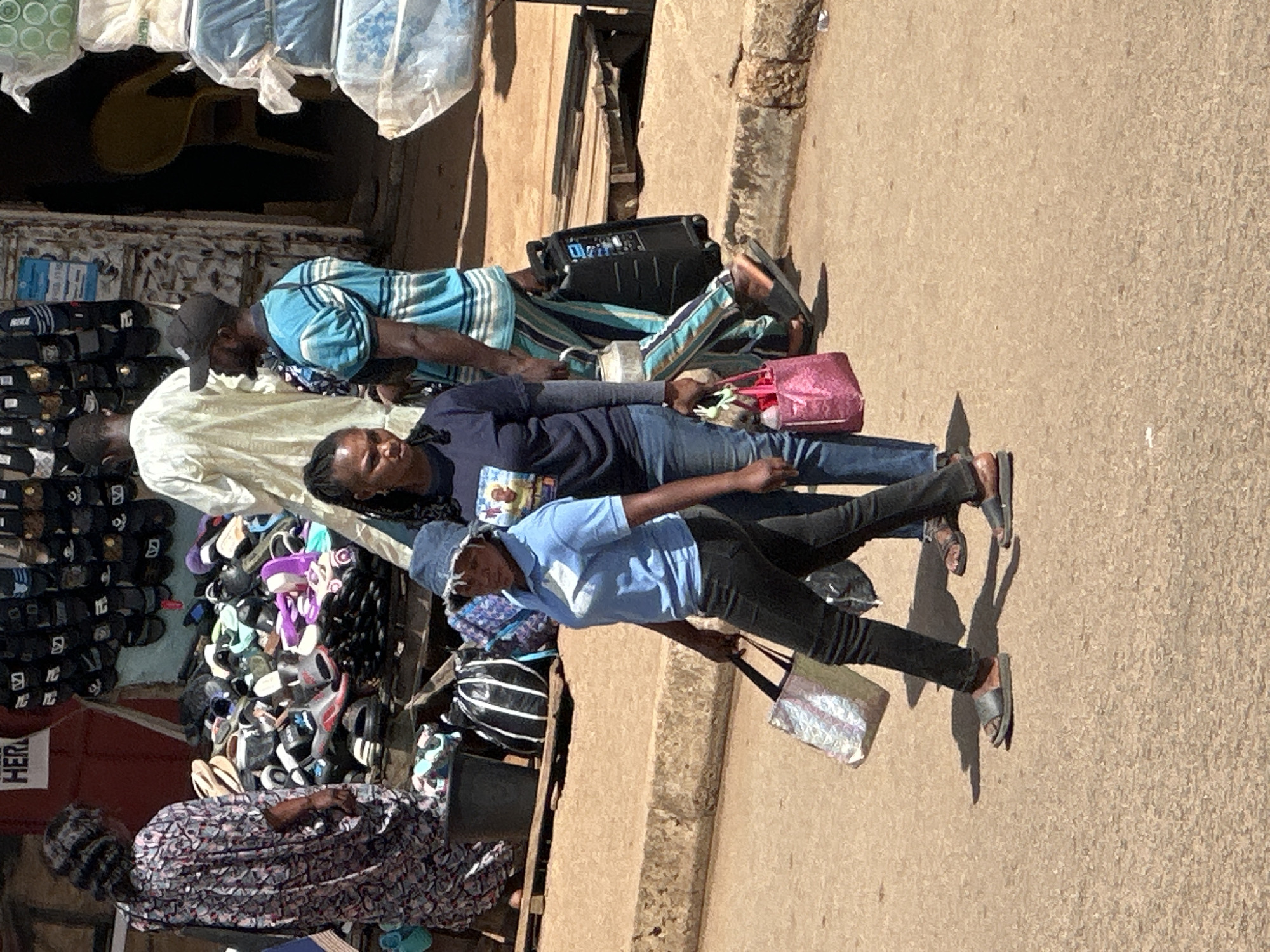 In Bukuru, Nigeria, an outdoor marketplace scene unfolds. A woman in a long, patterned dress stands to the left, facing a stall piled with footwear. To her right, a man wearing a light-colored robe is partially visible. In the foreground, two women walk across a paved surface. The woman on the left, wearing a blue shirt and denim hat, carries a patterned tote bag. The woman to her right, in a dark blue shirt, holds a pink tote bag and walks next to a man in a blue and white striped shirt and black cap, who is pulling a wheeled black case. In the background, a stall displays various flip-flops and sandals. The ground is a dusty, light brown, and a raised curb runs along the right side of the image.