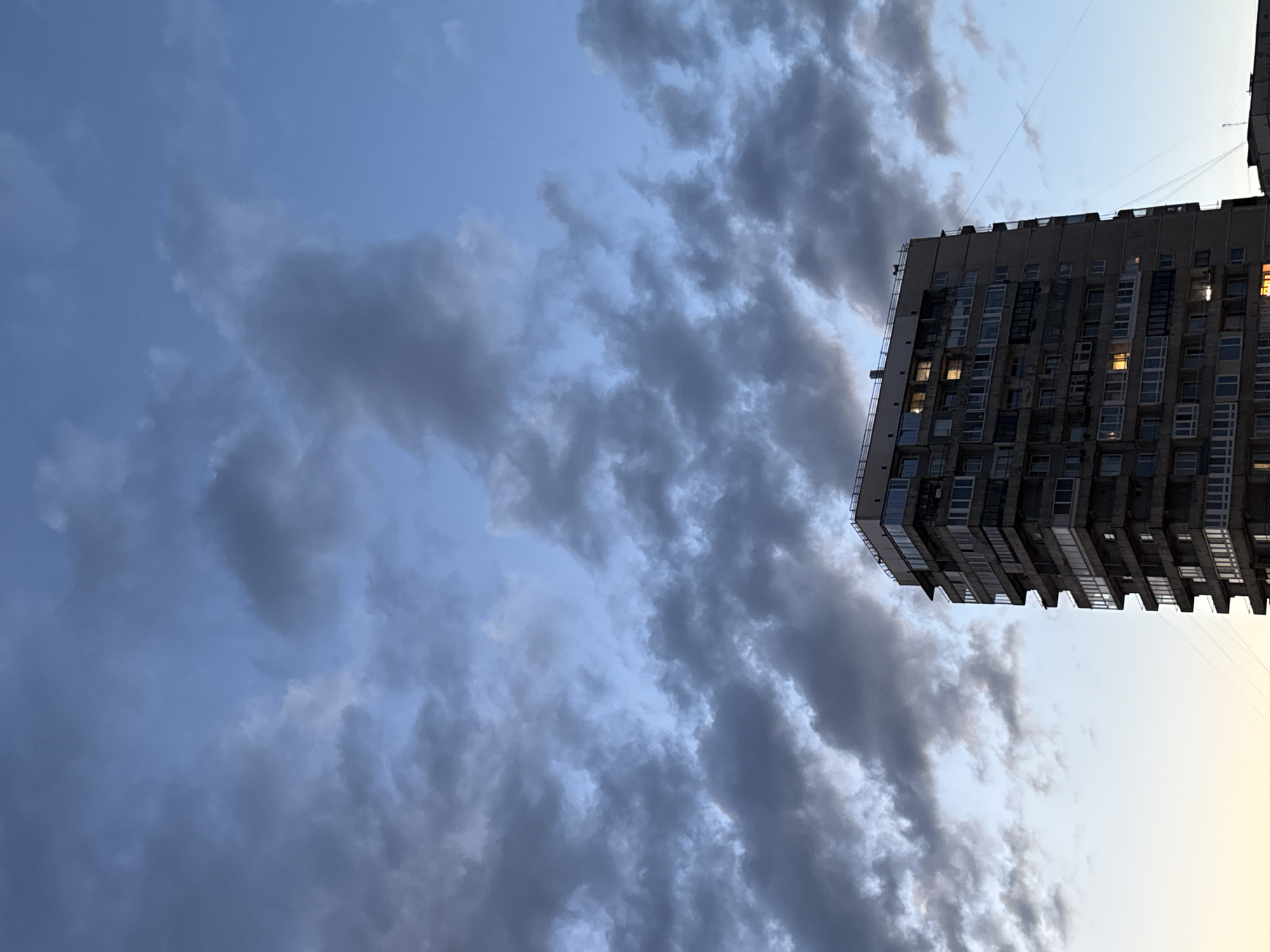 The image depicts a tall, multi-story building against a twilight sky filled with dark, dramatic clouds. The building appears to be made of concrete or similar material and has numerous windows, some of which are illuminated, suggesting occupancy. The architecture is typical of Soviet-era residential buildings. The sky is a gradient of deep blue and grey, with hints of a lighter hue at the horizon, indicating the fading daylight. The building occupies the lower right portion of the frame, with the sky dominating the upper two-thirds. The scene suggests an urban environment, possibly a residential district in Saint Petersburg, Russia, characterized by its imposing and somewhat stark architecture under a moody sky.