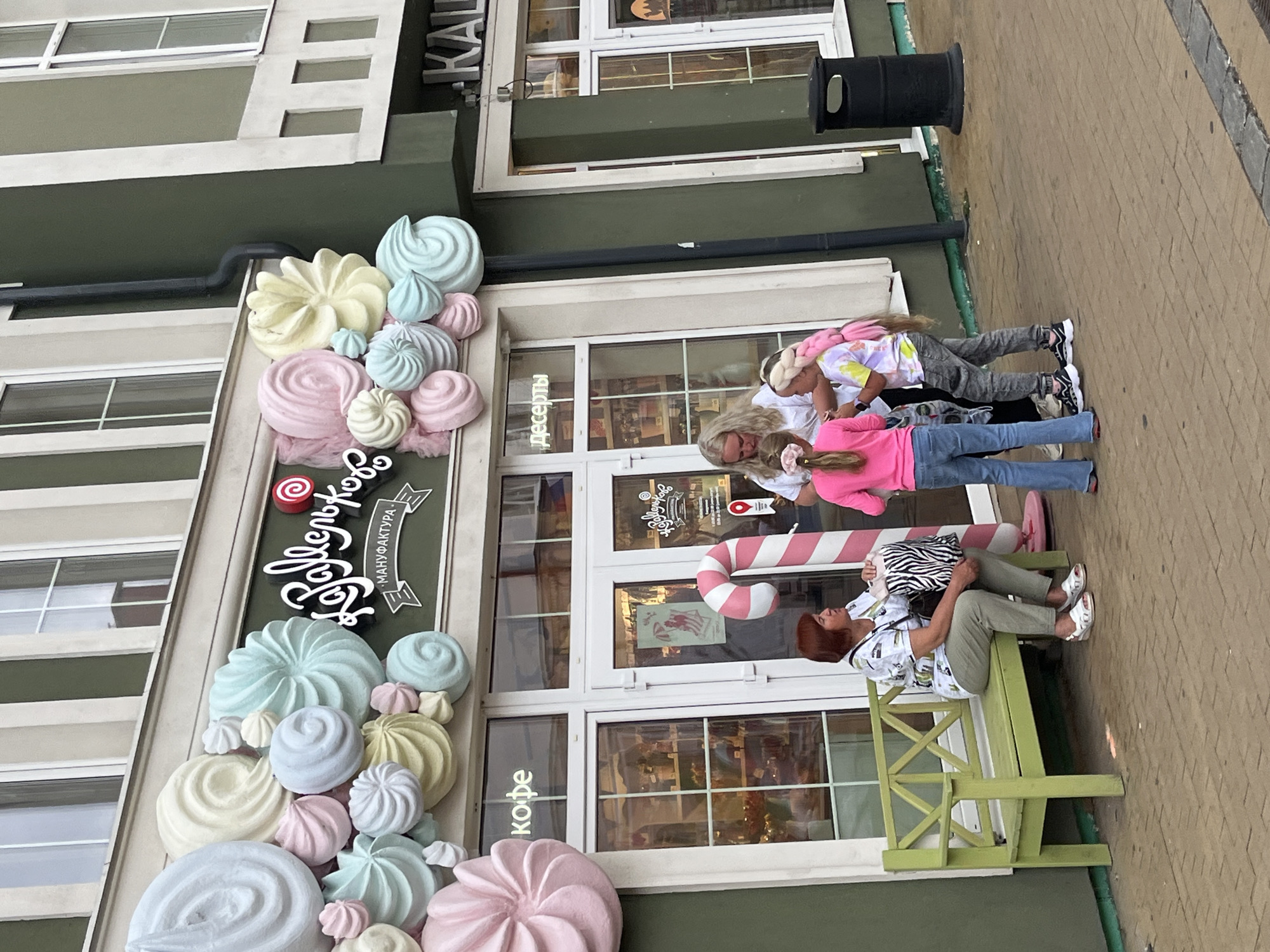 A storefront with large pastel meringue decorations on its facade advertises "Сладкоежка" (Sweet Tooth) and "Мануфактура" (Manufactory). The store features large windows displaying merchandise, possibly confectionery. A large pink and white candy cane prop stands near the entrance. Three people are visible outside the store. A woman with reddish-brown hair sits on a green park bench, holding a zebra-patterned bag. Two young girls, one with blonde hair and another with long pink braided hair, stand in front of the store entrance, interacting with a woman who appears to be a shop employee or a parent. The setting is outdoors, on a paved walkway in front of the building. The overall impression is that of a charming confectionery shop in a city, with elements suggesting a playful and sweet atmosphere. Based on the architectural style and the visible text, this scene is consistent with locations in Zelenogradsk, Russia, a resort town known for its picturesque streets and cafes.