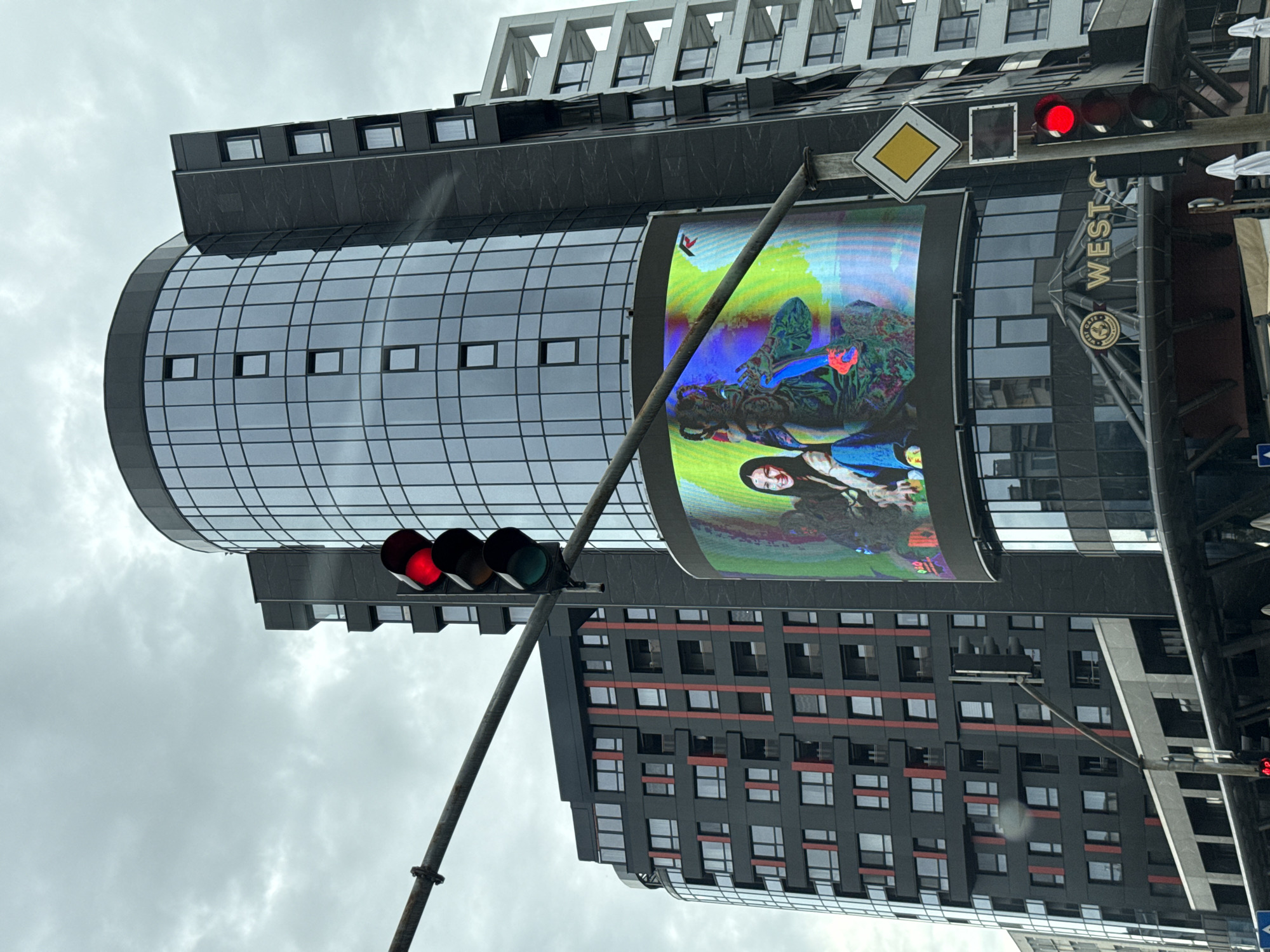 This image captures a street-level view in Plovdiv, Bulgaria, focusing on a modern, multi-story building with a prominent curved facade clad in dark glass. A large, vibrant digital billboard is integrated into the building's lower section, displaying a colorful image of two individuals, one appearing to be a woman with dark hair and the other a figure adorned in ornate, brightly colored clothing.
In the foreground, a traffic light is visible, with its red light illuminated, indicating a stop. A metallic pole supports the traffic light and a diamond-shaped yellow road sign with a black border, likely indicating a priority road. Behind the digital billboard, another building with a dark facade and red vertical accents is partially visible. To the right of the main building, a white, multi-story structure with a geometric, open-frame design is also in view.
The sky is overcast with a diffused light, suggesting a cloudy day. The overall scene depicts an urban environment with contemporary architecture and traffic control systems. The digital billboard is a focal point, adding a splash of color and visual interest to the somewhat muted tones of the surrounding buildings and sky.