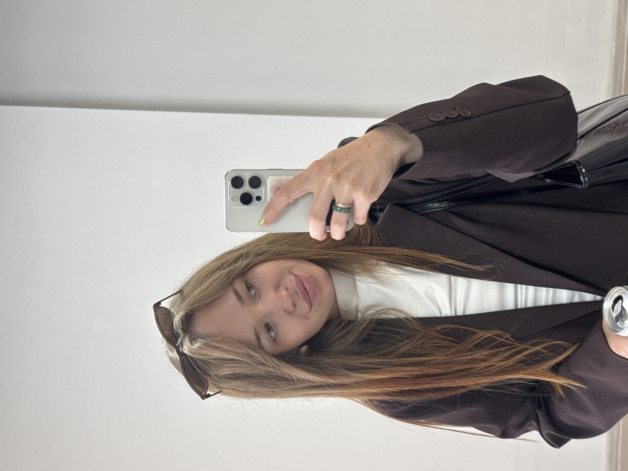 The image depicts a young adult female taking a mirror selfie in an indoor setting. The specific location in the city of Chaikovsky, Russia, cannot be confirmed from visual information within the image.
The woman has long, straight, light brown hair with reddish undertones, and sunglasses are perched on top of her head. She is looking to her right (viewer's left), towards the smartphone she holds, with a slight smile on her face. Her left hand (viewer's right) holds a silver-colored smartphone with three camera lenses, indicative of a modern model, and her index finger displays a light green nail polish. A small sticker is affixed to the back of the phone. Two rings, one green and one metallic, are worn on her left hand.
She is dressed in a white high-neck top beneath a dark brown blazer. The right sleeve of the blazer (viewer's left) shows three matching brown buttons on the cuff. Over her left shoulder (viewer's right), a portion of a black, possibly glossy, handbag is visible. Her right hand (viewer's left), partially visible at the bottom of the frame, holds a silver beverage can.
The background consists of plain white walls, with a distinct vertical seam or corner visible on the right side of the frame, suggesting an interior room. The lighting is bright and even, casting minimal shadows.