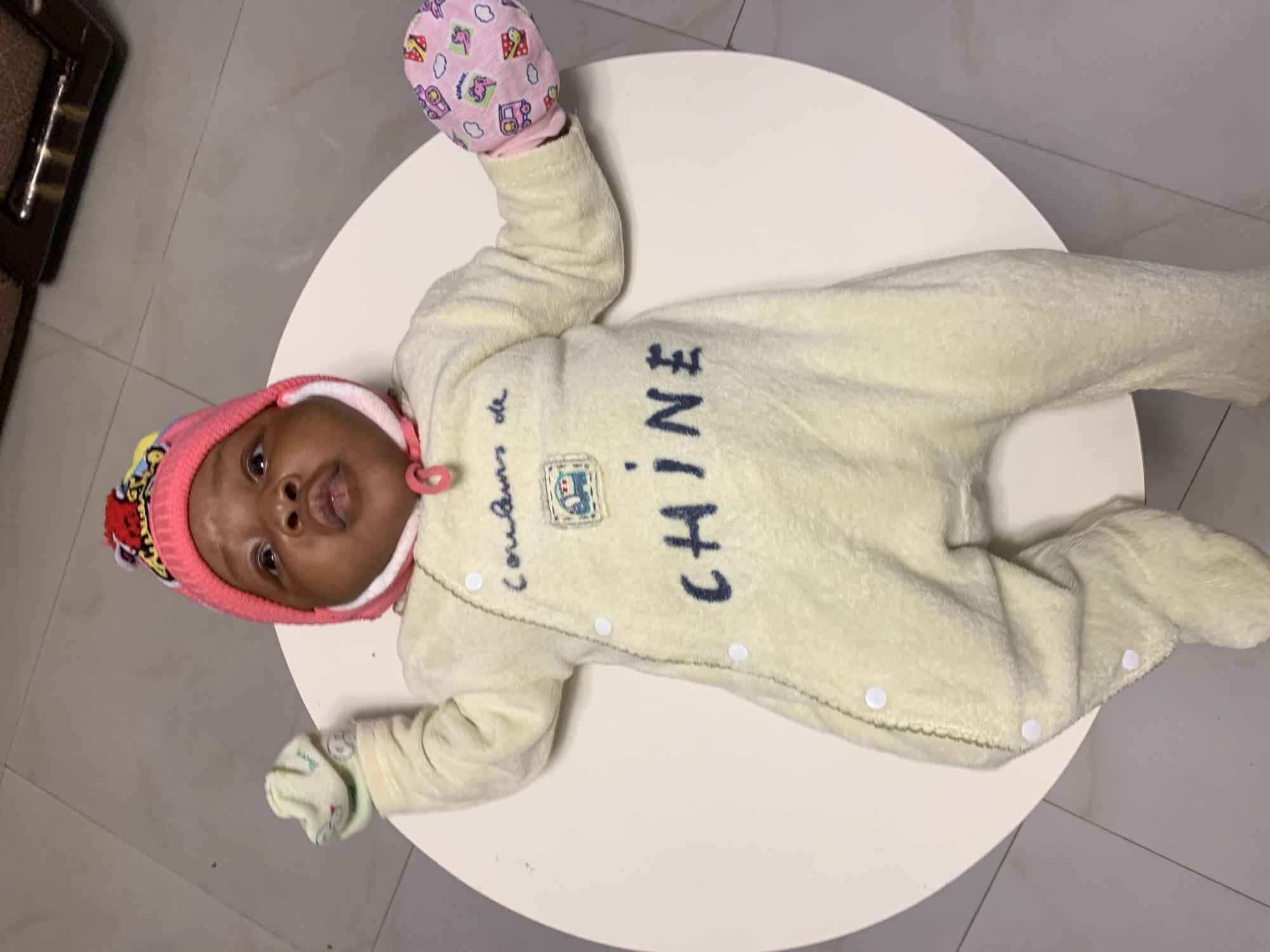 An infant is depicted lying on their back on a round white table, dressed in warm attire. The baby has dark skin and is wearing a pink knitted hat with colorful patterns, and a light yellow or cream-colored full-body jumpsuit. On the baby's left hand (viewer's right) is a pink mitten with colorful patterns, and on the right hand (viewer's left) is a light green mitten, also with patterns. The jumpsuit features white snap buttons running down the front. Visible text on the jumpsuit reads "Couleurs de CHINE," with "Couleurs de" in a smaller, cursive script and "CHINE" in larger, block letters. A small embroidered patch with an image is present below "Couleurs de." The baby's head is tilted slightly, and they are looking towards the camera with a slightly open mouth and a neutral, curious expression. The background consists of a gray tiled floor, and in the upper left corner, part of a dark brown furniture piece is visible. The scene suggests an indoor setting, likely a home in Rafin Bauna, Nigeria. The overall impression is one of a comfortable and calm baby.